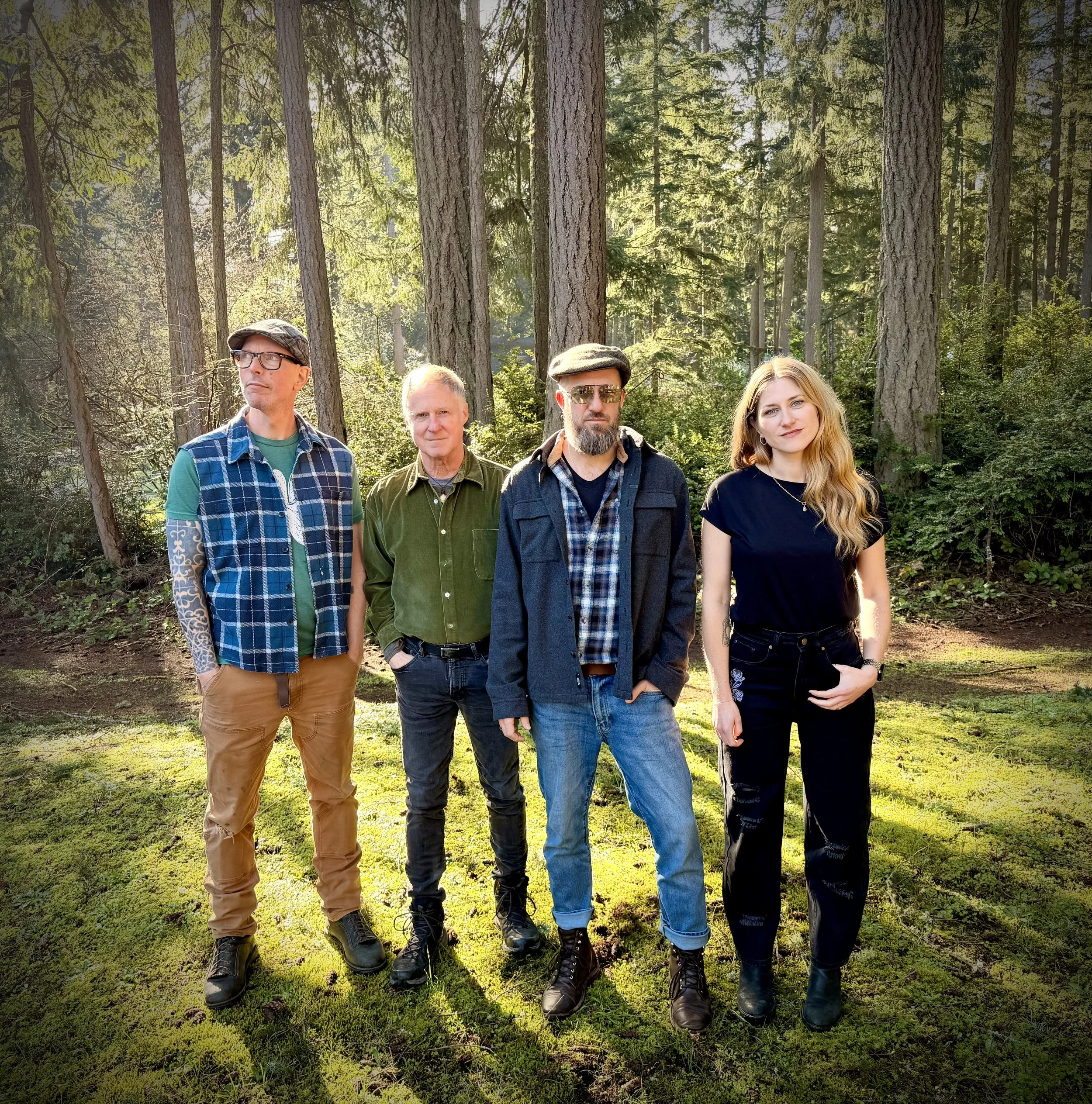 Four people standing outdoors in a forest, posing for a photo. The group includes three men and one woman, all dressed casually and standing on a grassy area with tall trees in the background.