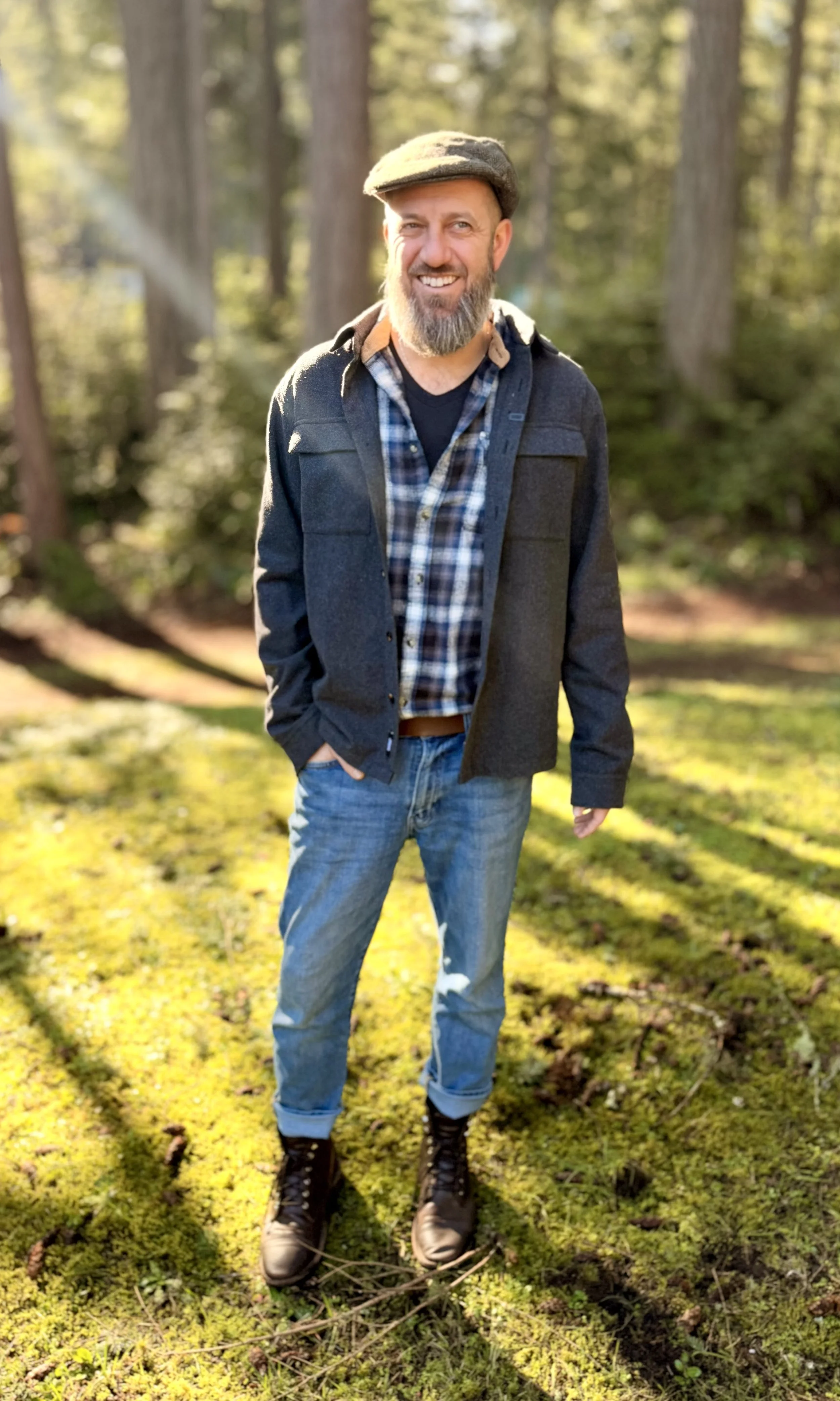 A man with a beard and mustache, wearing a flat cap, plaid shirt, dark jacket, jeans, and boots, standing outdoors in a forest with tall trees and sunlight filtering through the leaves.