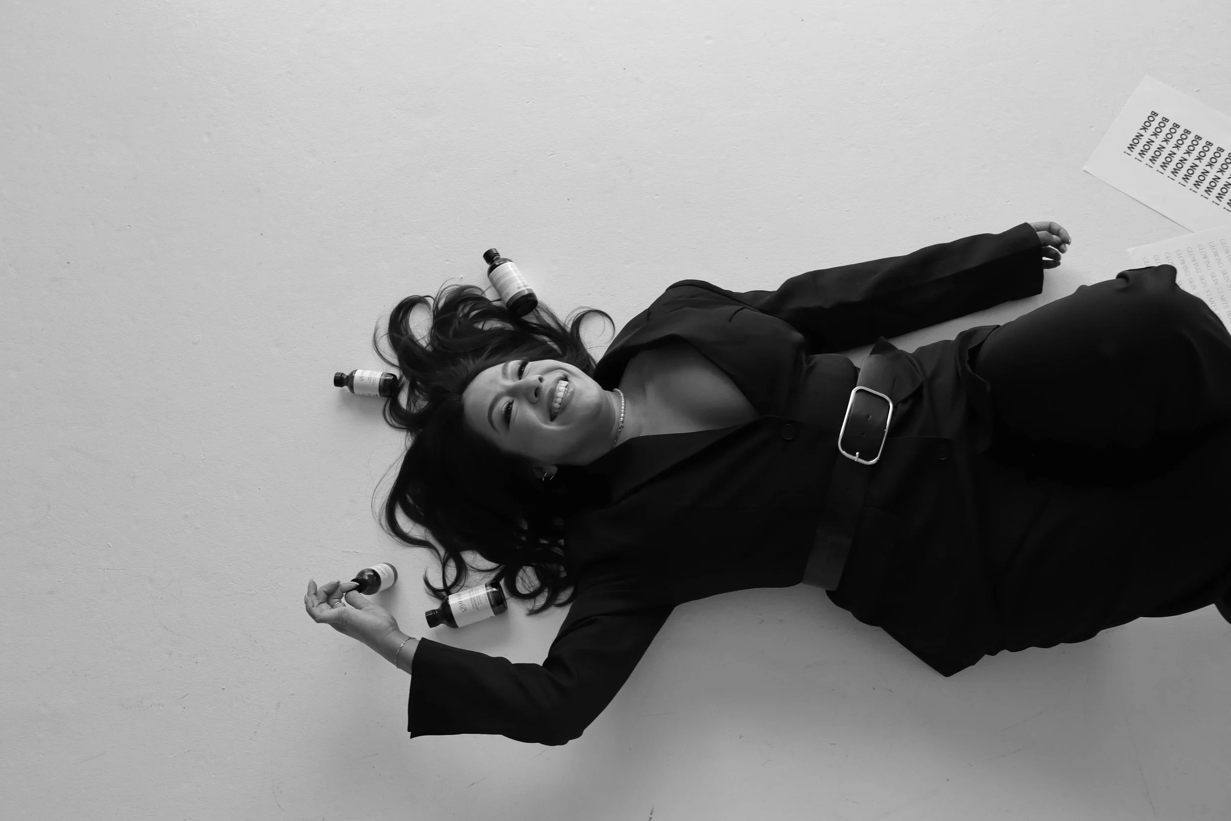 A woman lying on her back on a plain white floor, smiling, with long wavy hair. She is dressed in black clothing and surrounded by several small bottles or jars, some of which are placed on her hair and body. There is a piece of paper or card nearby with text on it.