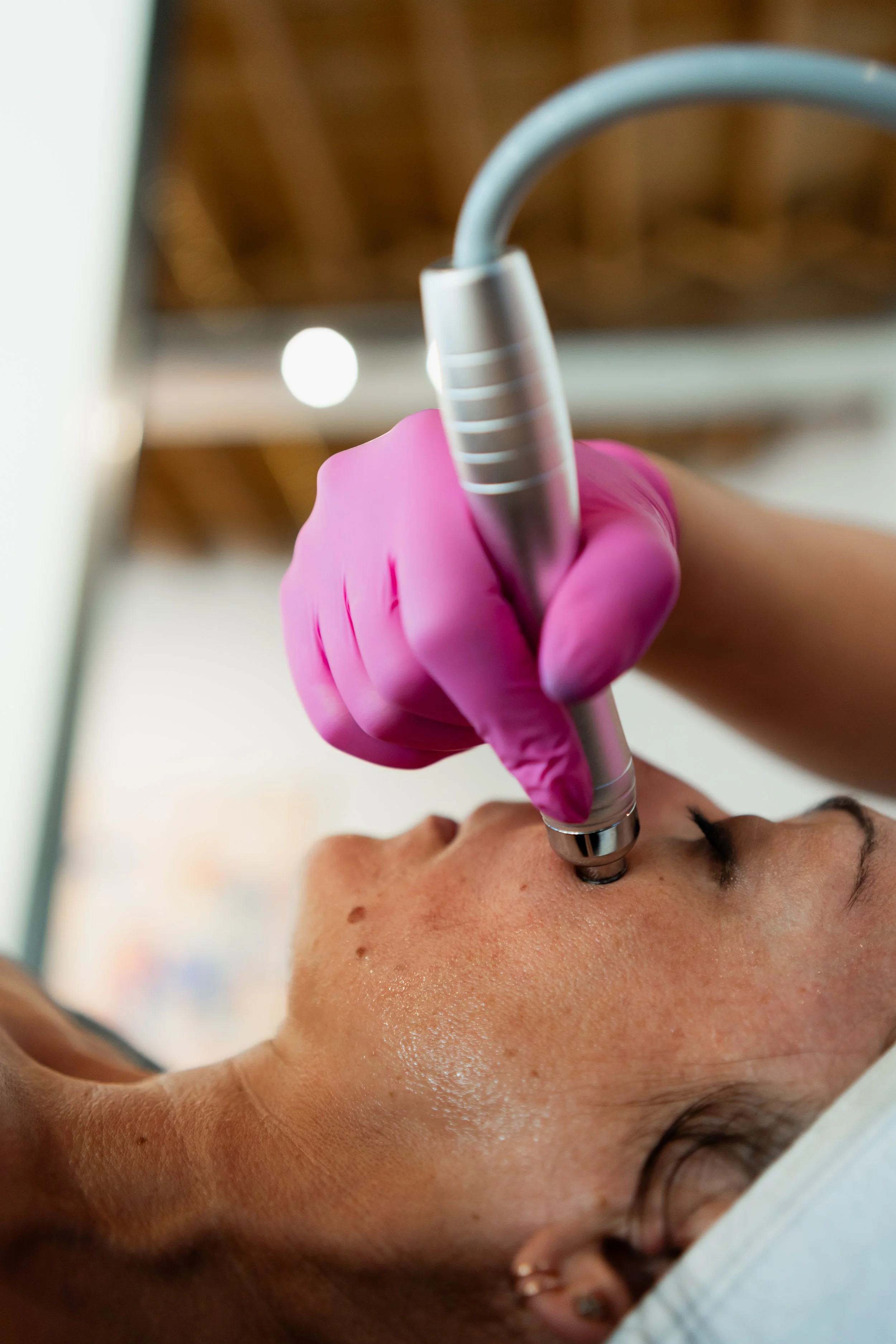 A person receiving a facial treatment with a handheld device on their face, wearing a pink glove, in a clinical setting.