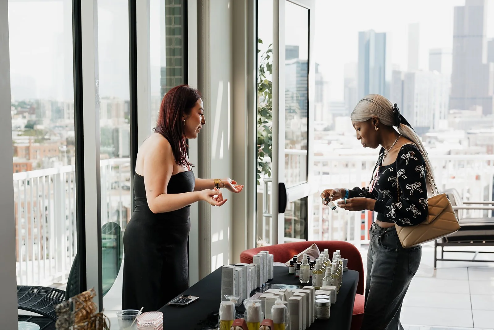 Two women standing at a table with skincare products on display in a high-rise building with large windows and a city view.