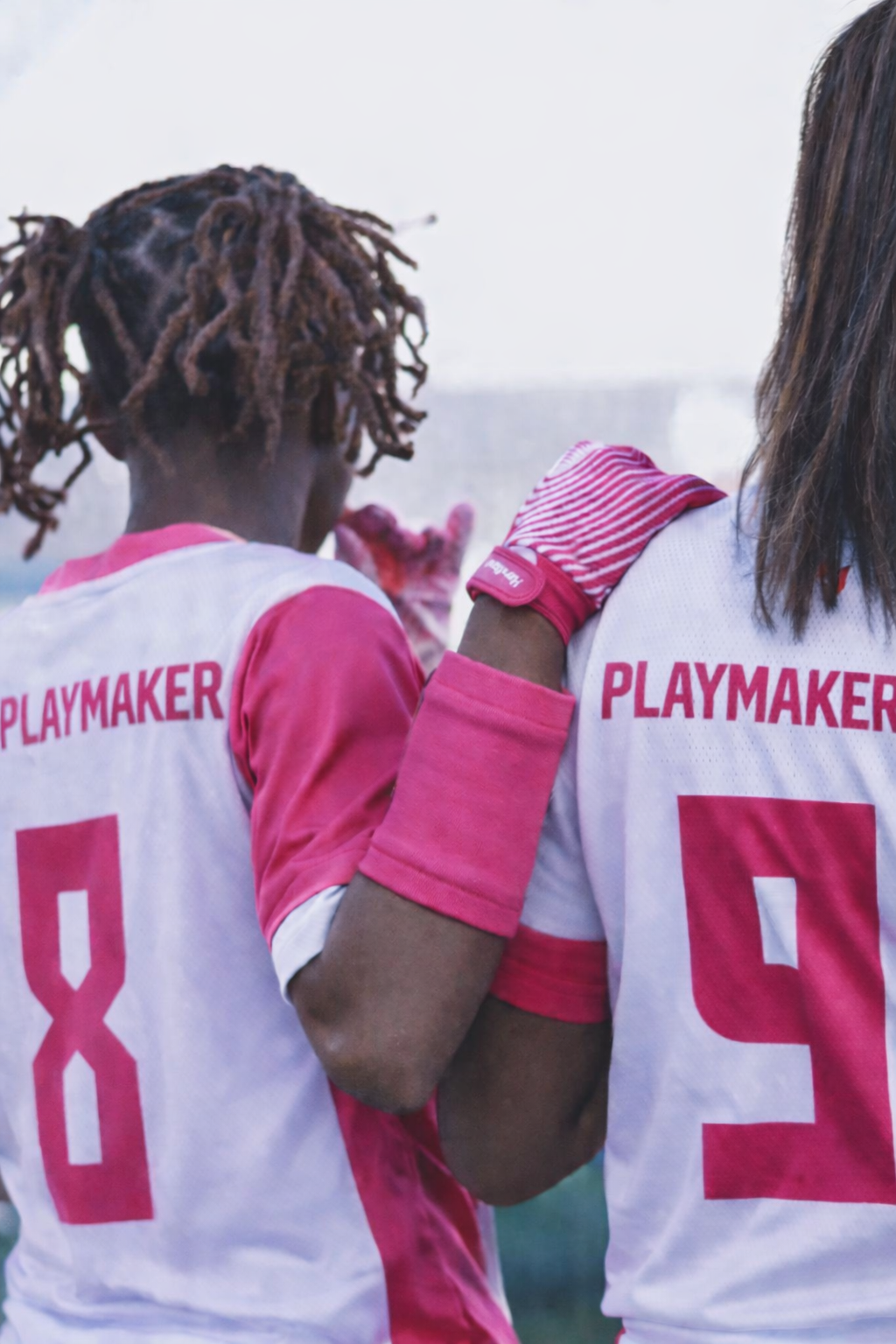 Two female football players standing together on the field, wearing white and pink jerseys with the word "PLAYMAKER" on the back and numbers 8 and 9. One has dreadlocks, and the other has long straight hair, both with pink accessories.