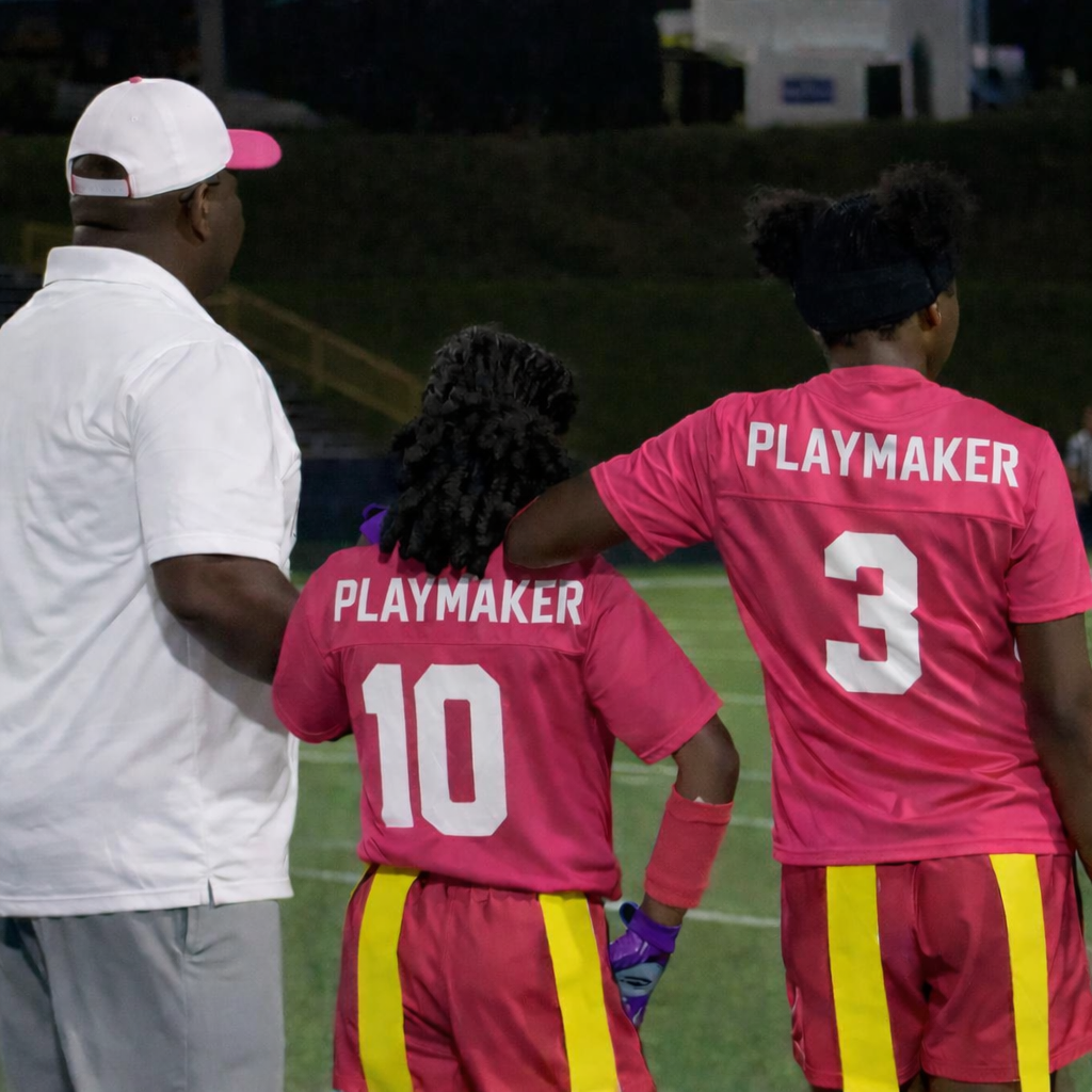 Two female athletes with dark skin in pink uniforms with 'PLAYMAKER' on the back, numbered 10 and 3, standing on a football field with a coach during a game or practice, the player with number 3 has her arm around the player with number 10.