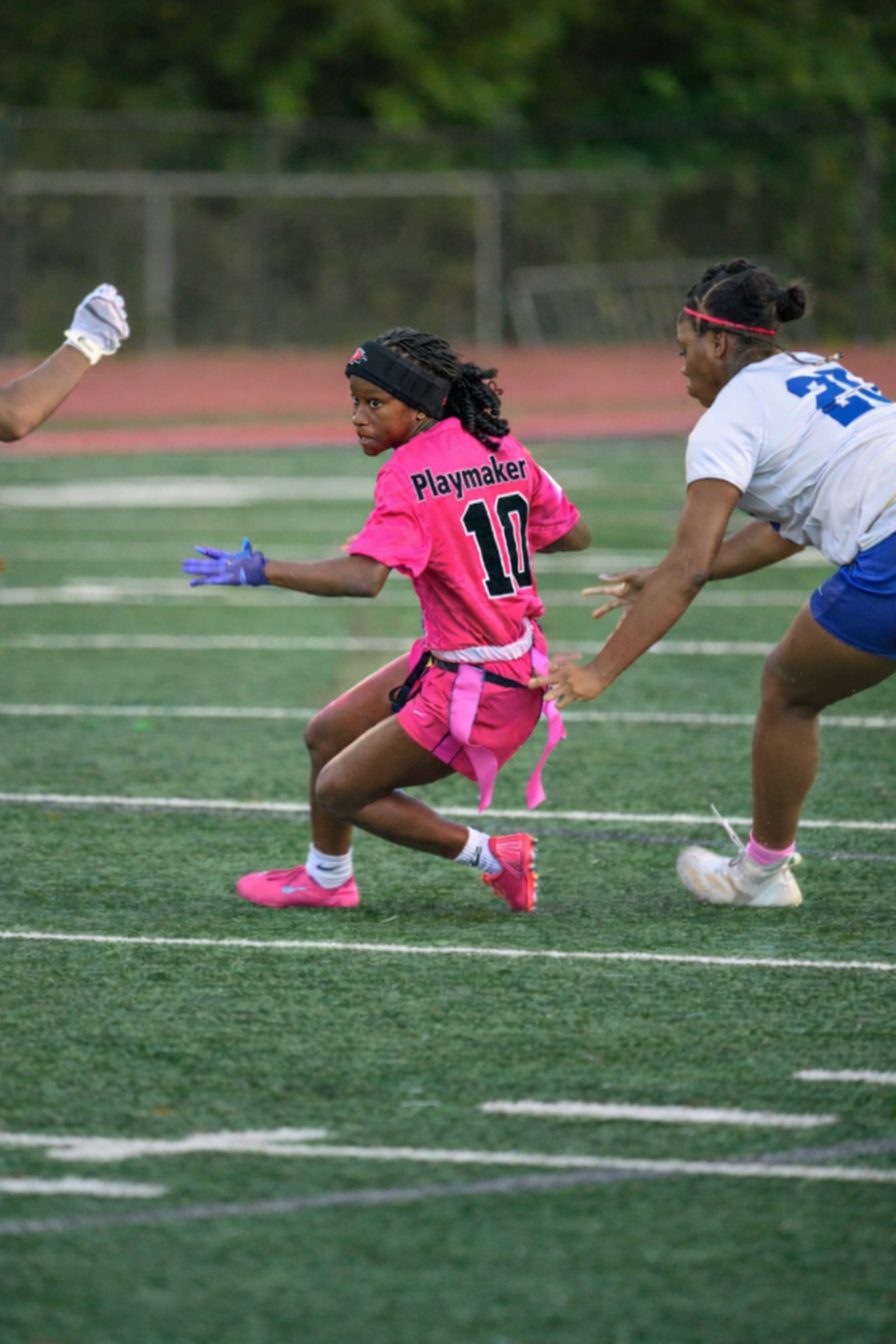 Two girls playing flag football on a football field. The girl in the foreground is wearing a bright pink jersey with the number 10 and the word 'Playmaker' on the back, running with a focused expression. The other girl, dressed in a white jersey with blue shorts, is reaching out to grab her flag.
