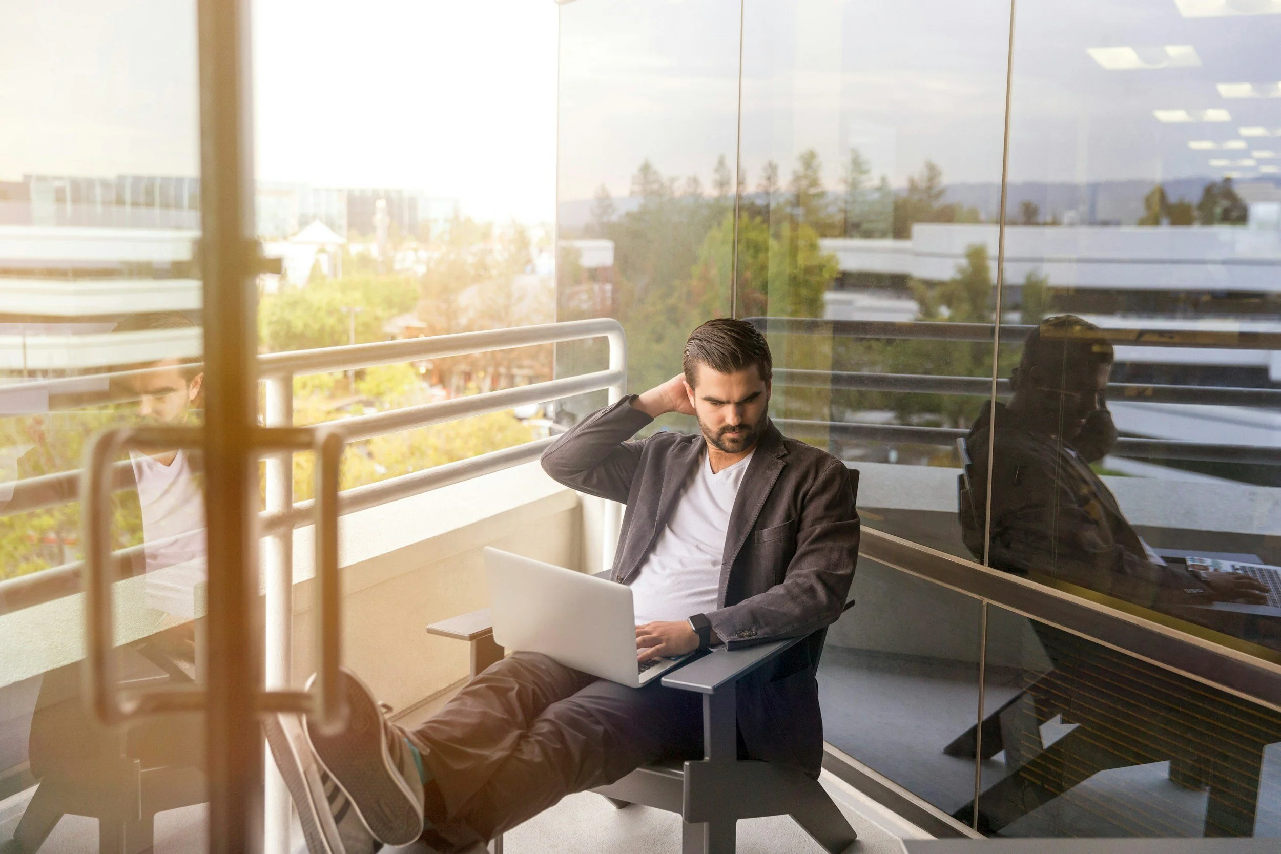 A young man sitting on an office balcony wearing a white t-shirt, brown sport coat, jeans and sneakers with a notebook computer in his lap.