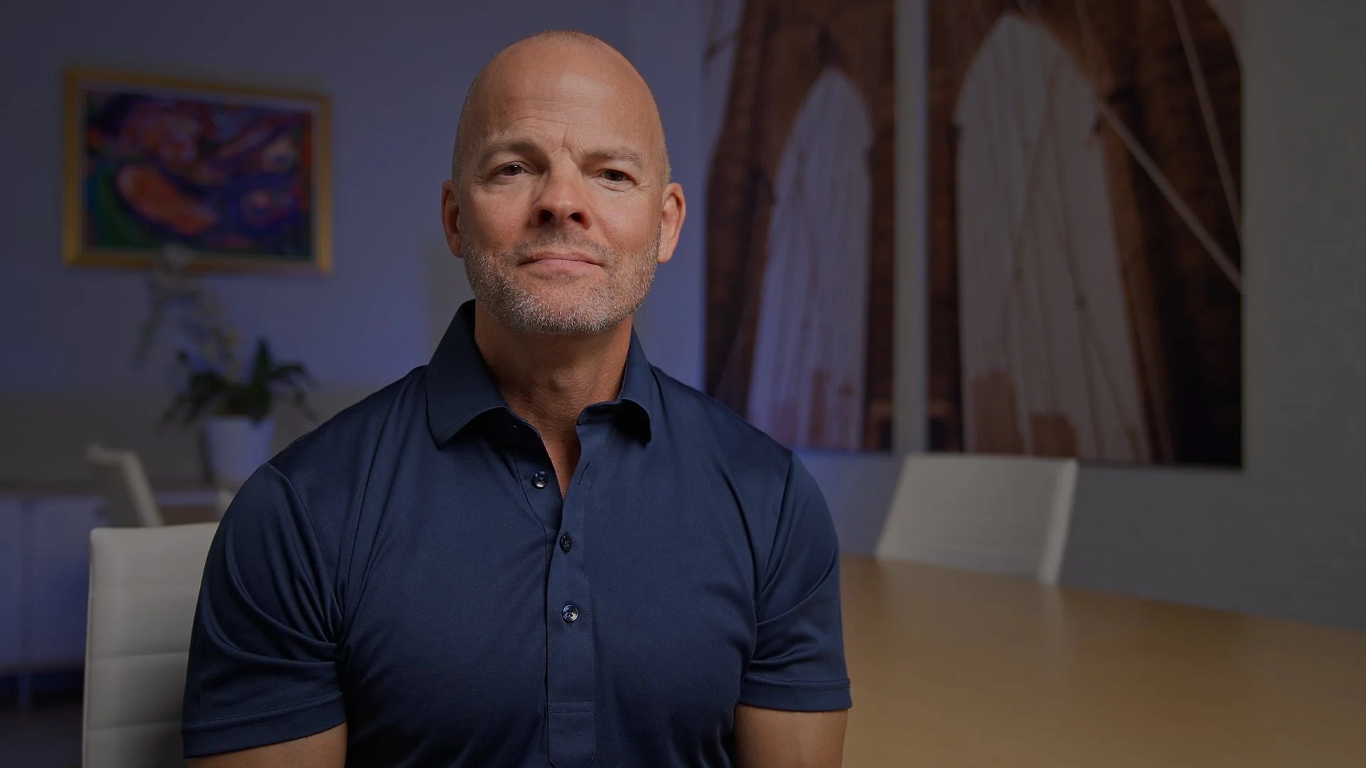 A middle-aged man with a bald head and beard, wearing a navy blue collared shirt, sitting in a modern conference room with abstract artwork on the wall behind him.