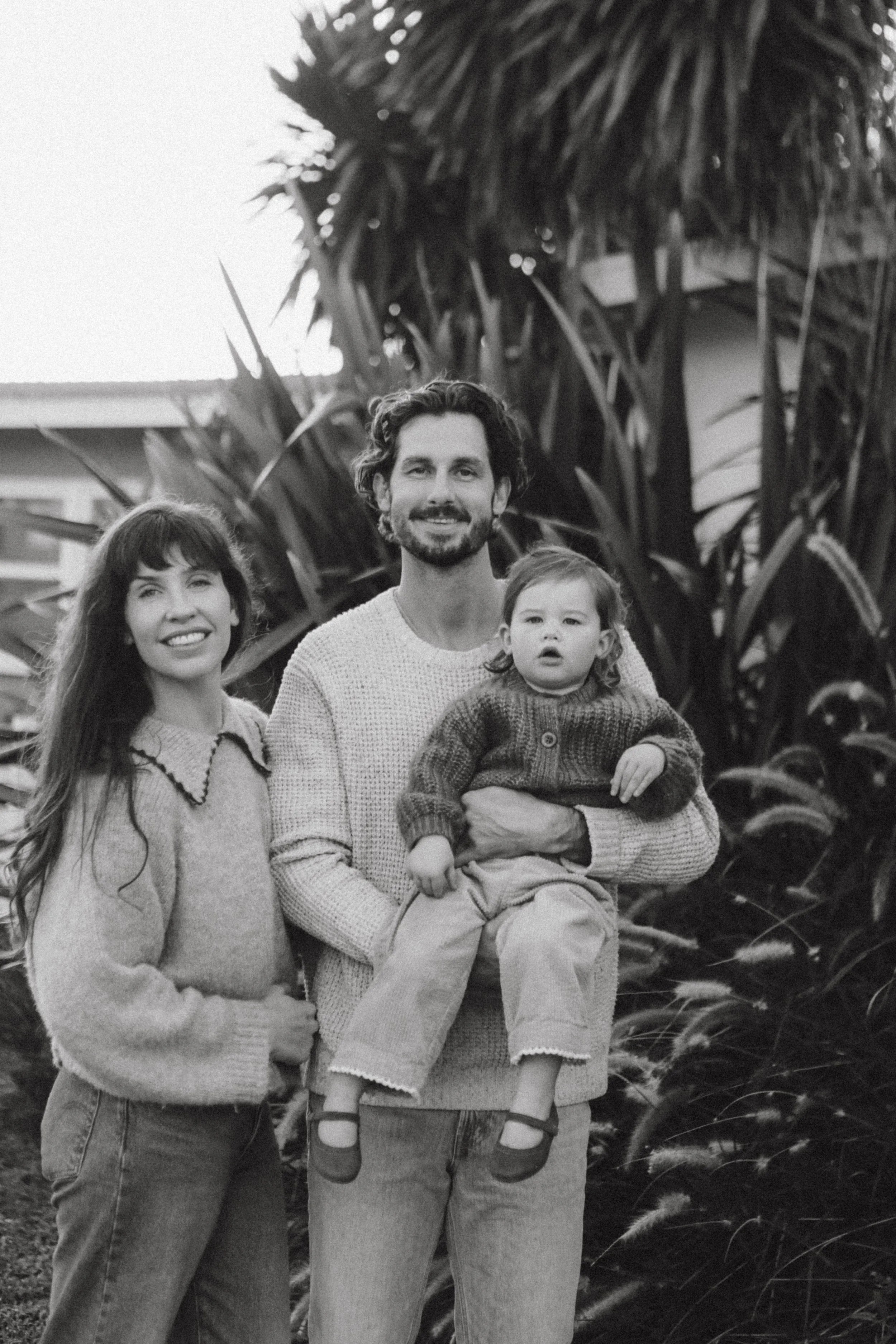 A black and white photo of a family of three standing outdoors in front of tall plants. The woman has long hair, is smiling, and wearing a sweater. The man has medium-length curly hair, a beard, and is holding a young girl who is wearing a sweater and pants. The girl looks curious, and the family appears happy.