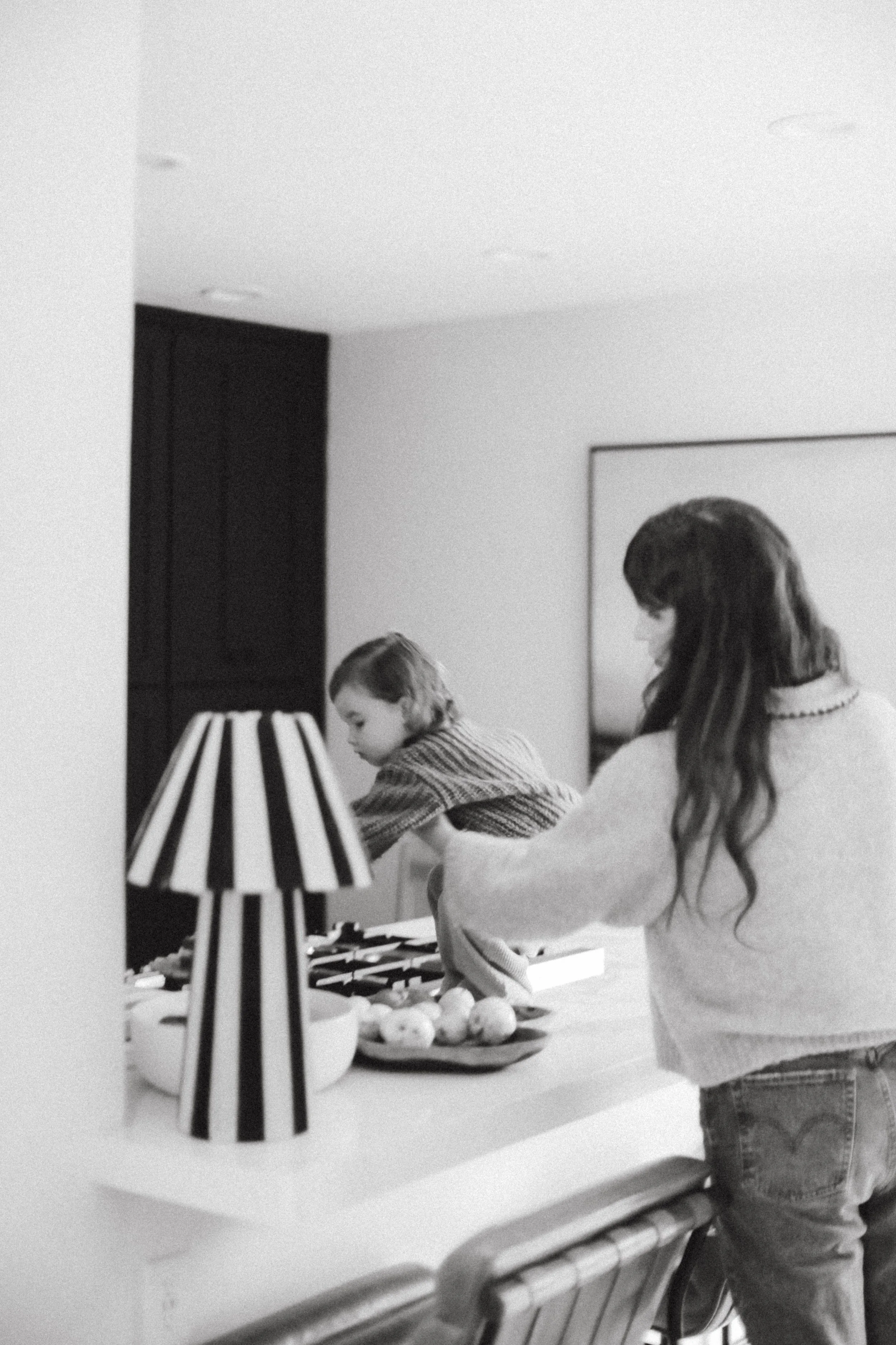 A woman and a child standing at a kitchen counter, preparing food or snacks, with a table lamp on the counter, black and white photo.