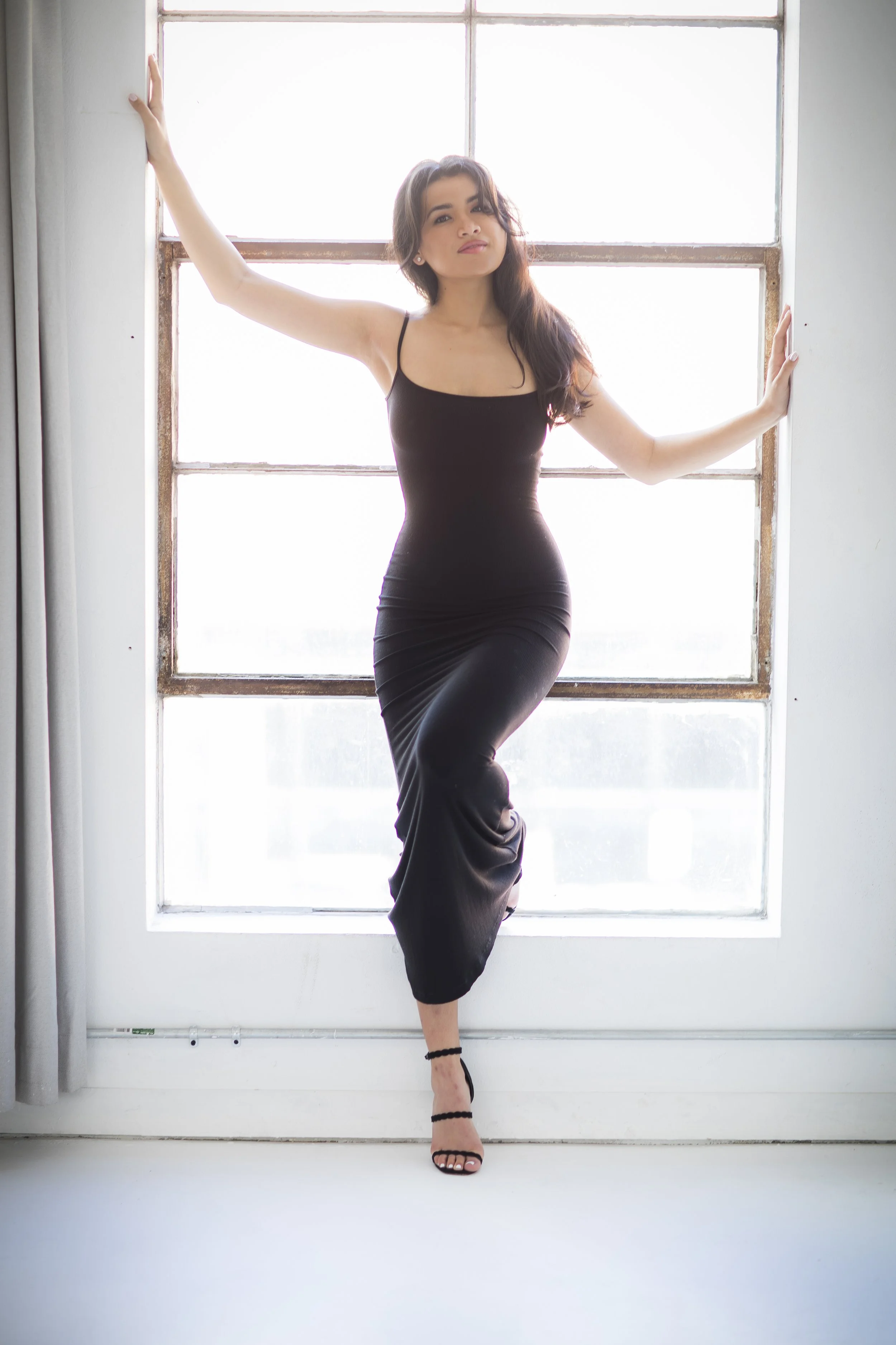 A woman in a black dress standing in front of a large window, with one hand on the wall and the other on the window frame.