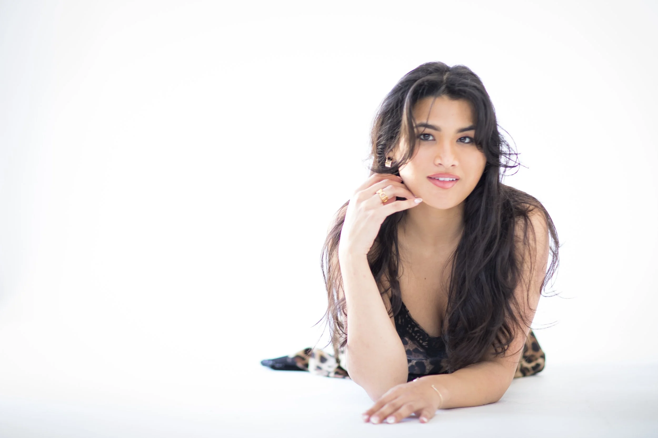 A woman with long dark hair, wearing a leopard print top, is lying on her stomach on a white background, resting her chin on her hand, looking at the camera with a soft smile.