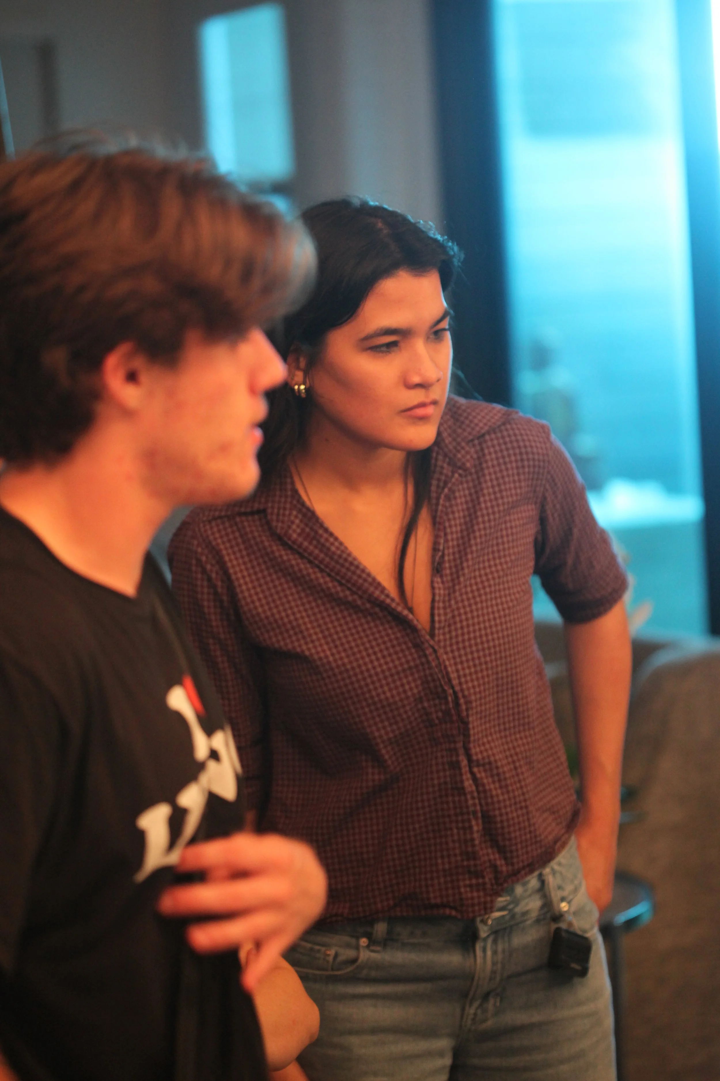 A woman with dark hair wearing a checkered shirt and jeans and a man with brown hair in a black shirt are standing indoors, engaged in a conversation, with blurred background of windows and furniture.