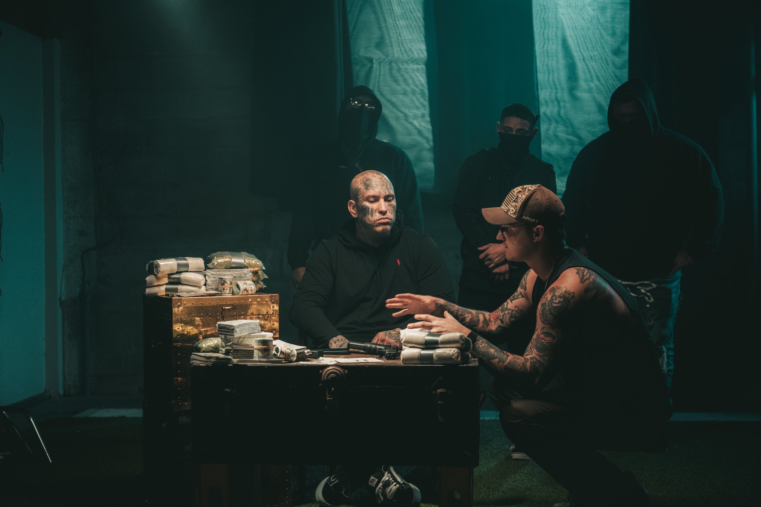 Group of men with tattoos discussing around a table stacked with large quantities of wrapped packages, some in gold and others in silver, in a dimly lit room.