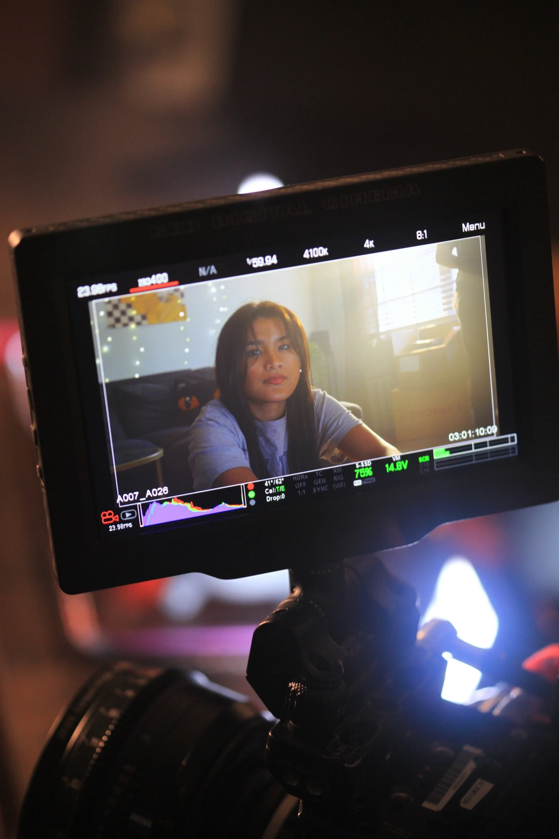 Close-up of a camera monitor showing a young woman with long dark hair sitting indoors by a window, looking at the camera with a neutral expression.