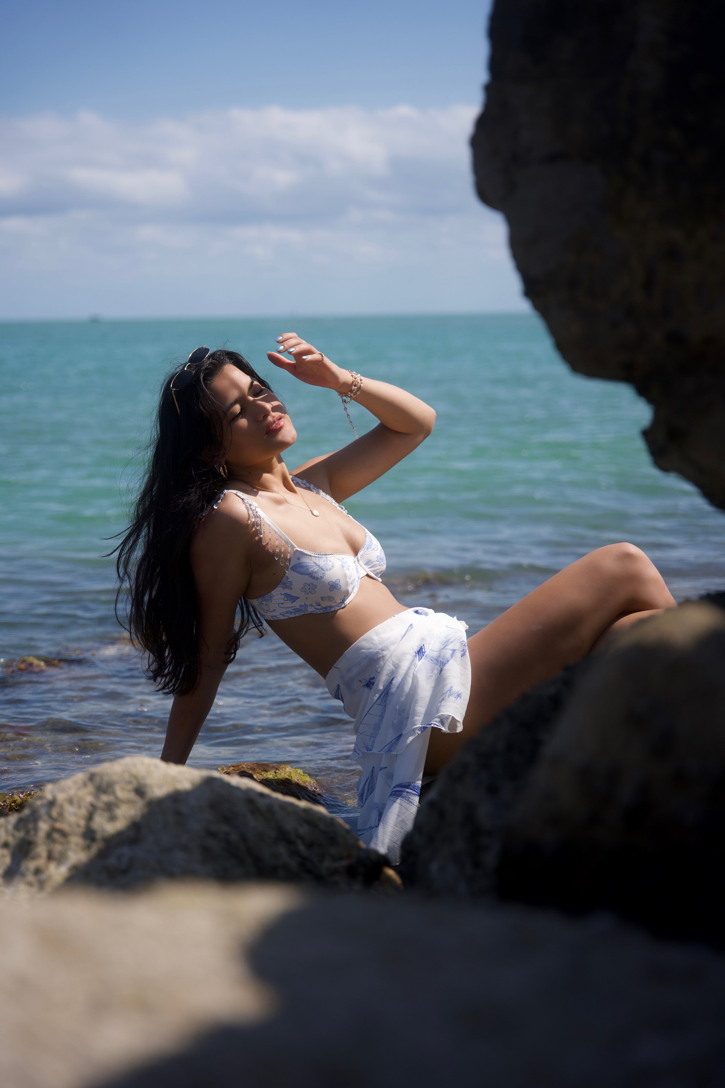 A woman in a floral bikini and matching skirt relaxing on rocks by the ocean, with her eyes closed and hand shading her face from the sun.
