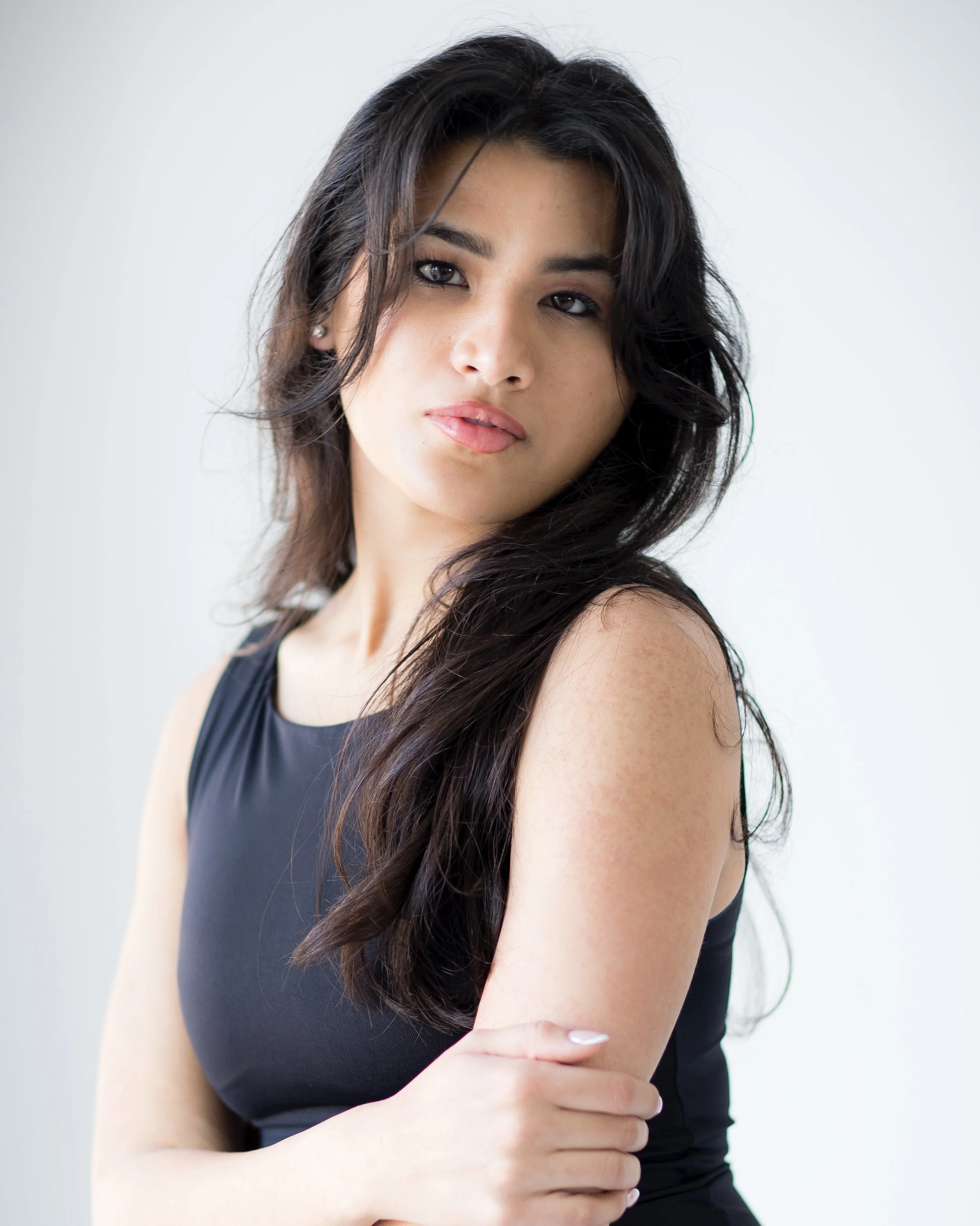 A young woman with long, dark hair wearing a sleeveless black top, posing against a plain white background.