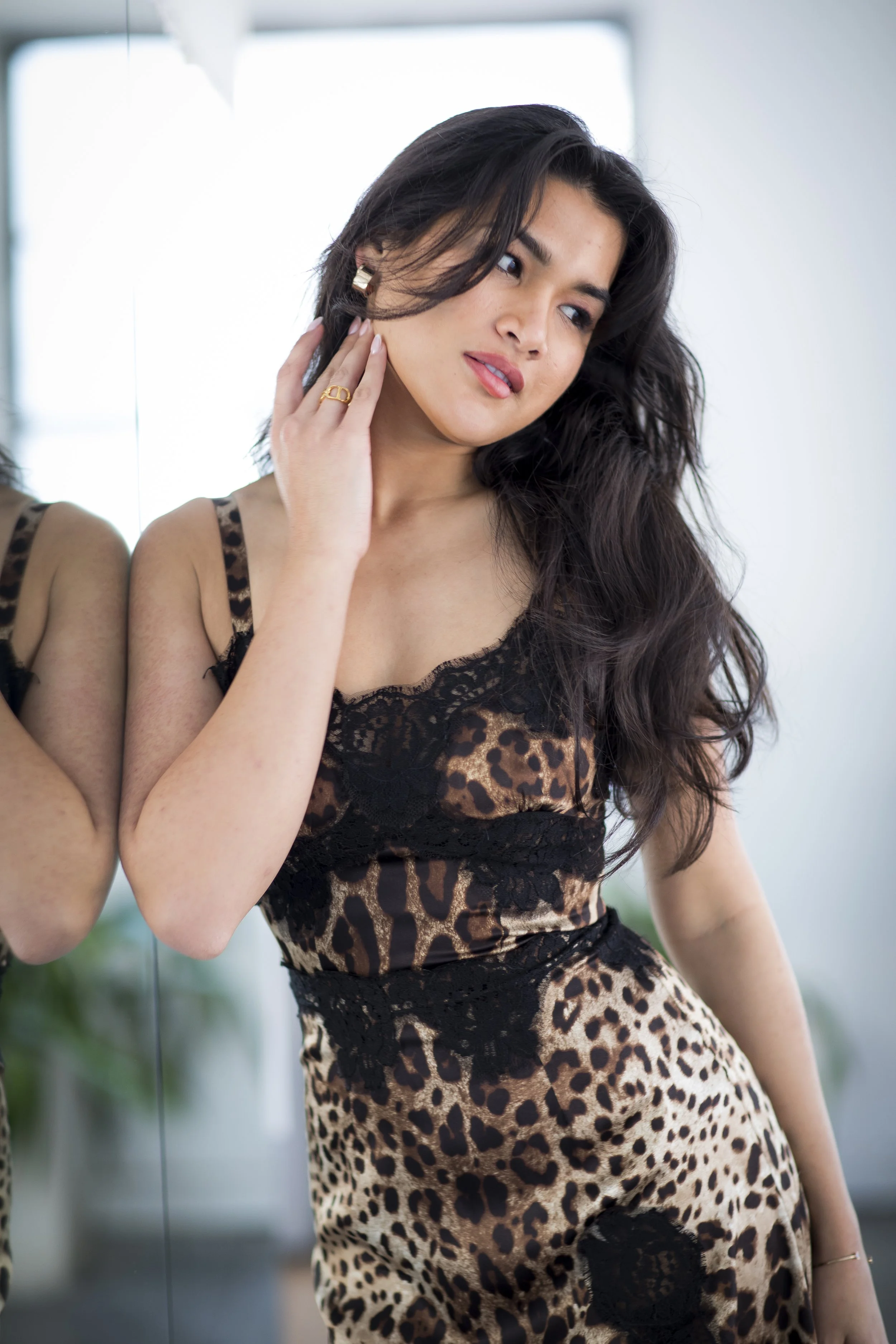 A woman with long dark hair wearing a leopard print dress with black lace details, standing in front of a mirror.