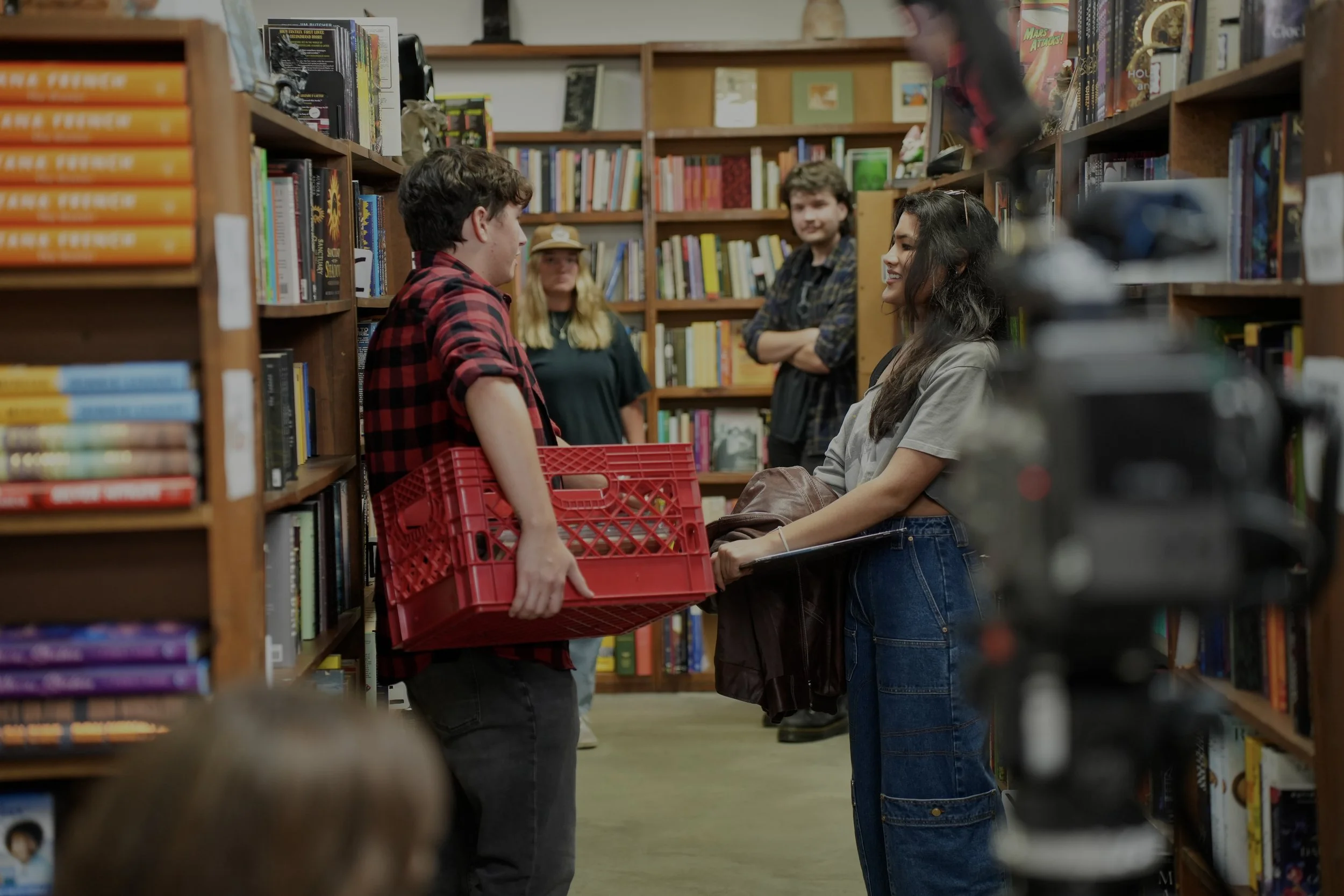 Two teenagers are standing opposite each other in a bookstore aisle, smiling and talking. The boy is wearing a red and black checkered shirt and holding a red plastic basket, while the girl is wearing a gray top and holding a brown bag. In the background, three other people are visible; a woman with long blonde hair and a hat, and two boys, one with folded arms, smiling at the conversation.