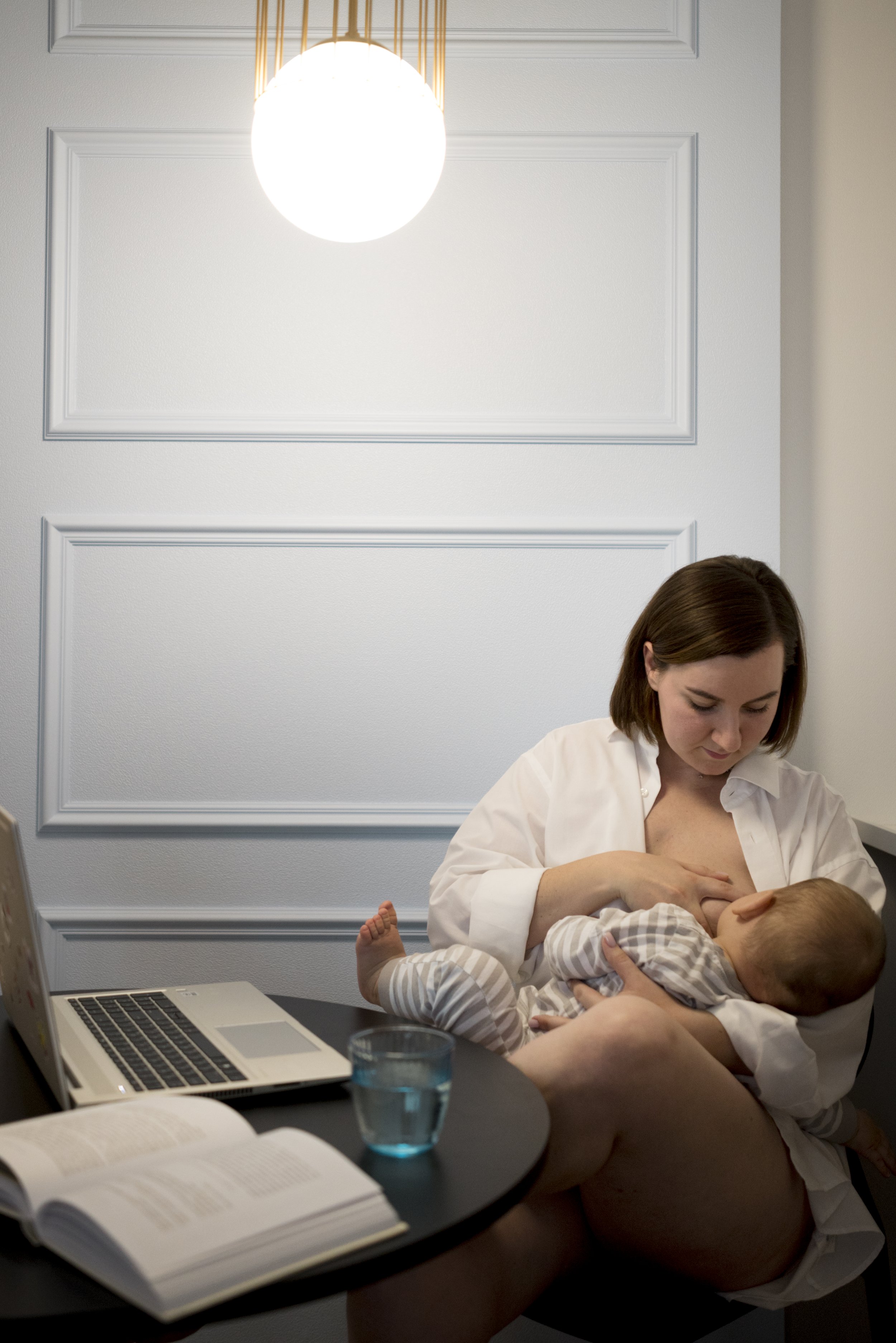 A woman breastfeeding a baby at a desk with a laptop, glass of water, and an open book against a white paneled wall with a bright ceiling light.