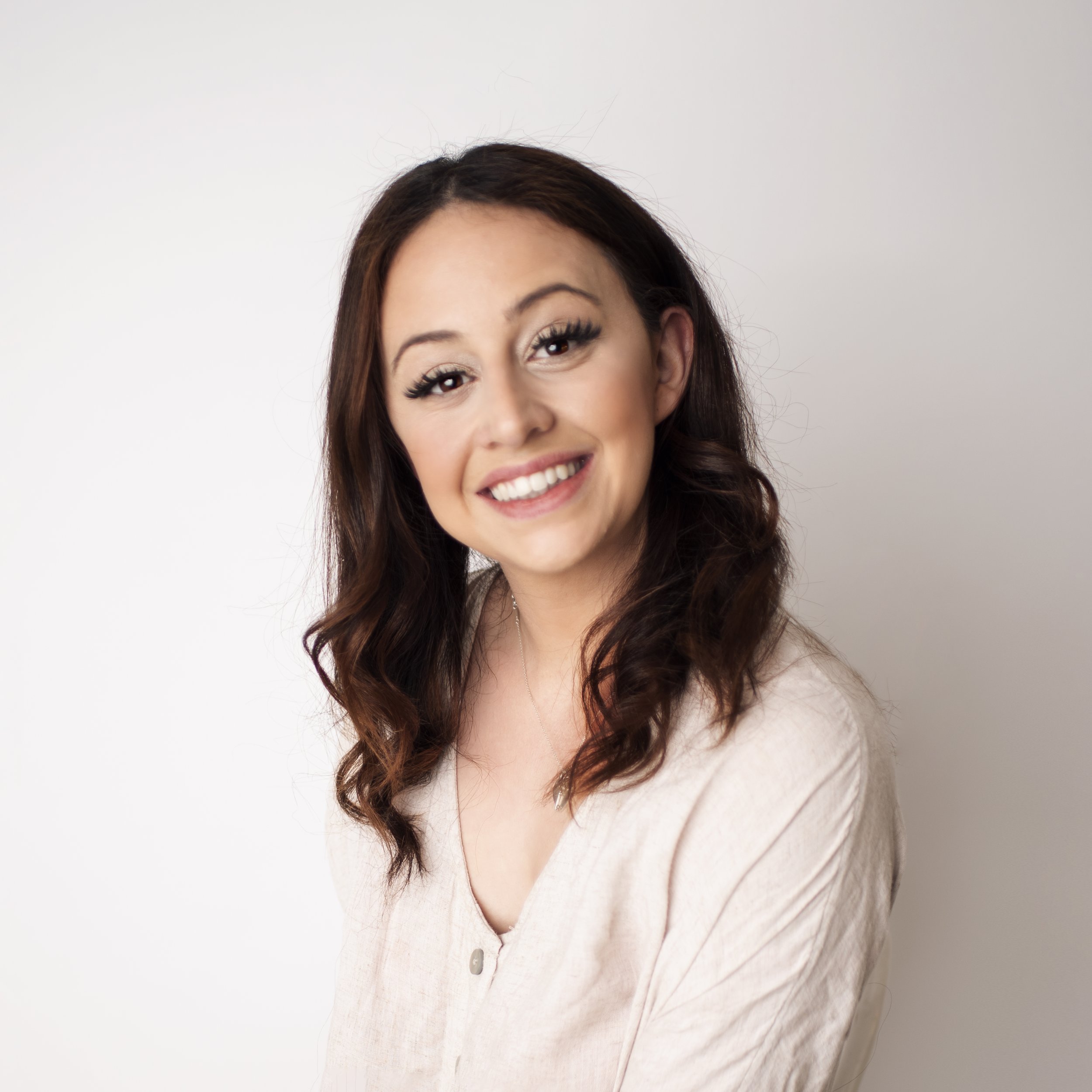 Portrait of a smiling woman with long, curly dark hair, wearing a beige top against a plain light-colored background.