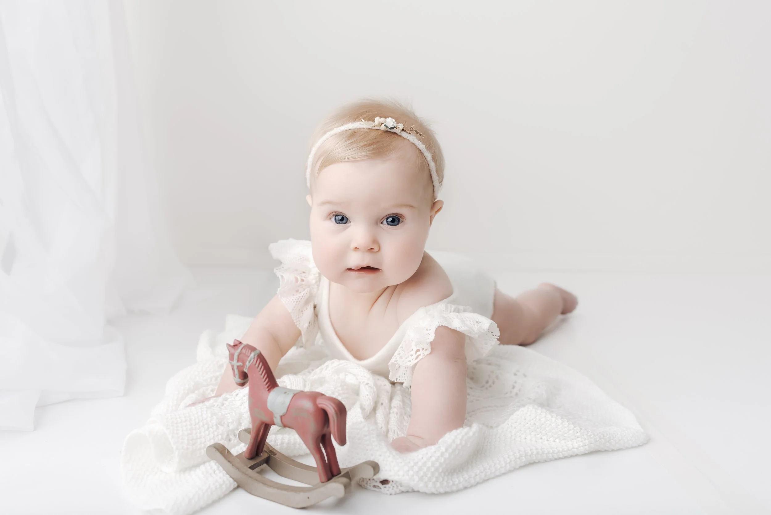 Baby girl with a headband, on stomach on a white blanket, with a small wooden rocking horse toy.