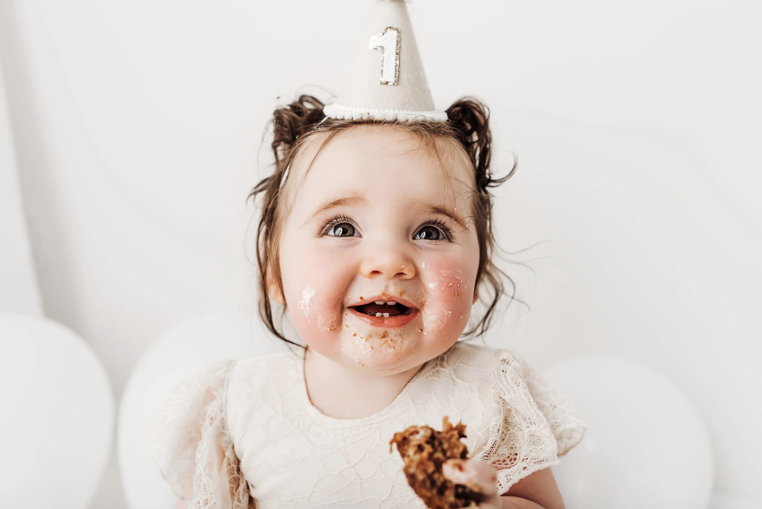 A young girl with curly hair wearing a birthday hat with a number one on it, smiling with cake and frosting on her face, holding a piece of cake.