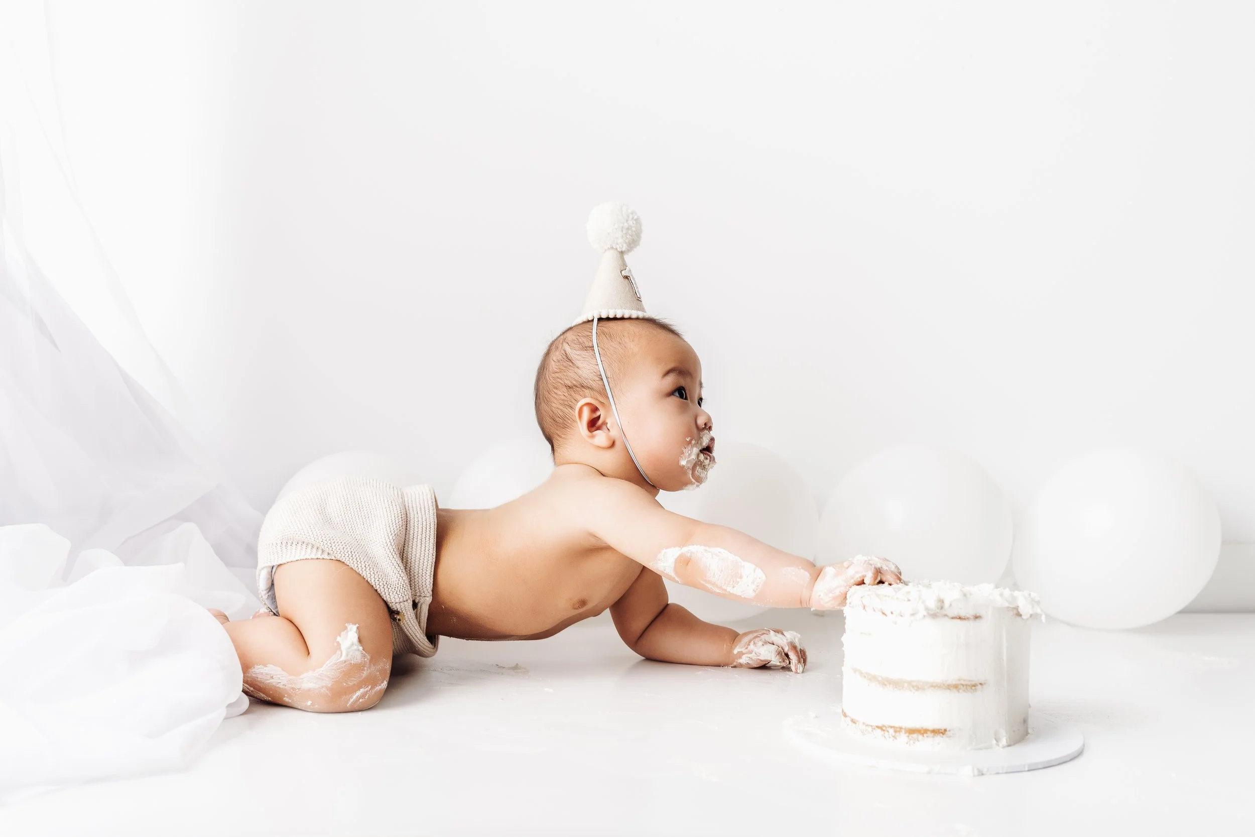 Baby in a beige party hat crawling on a white surface, reaching for a small white cake with a single candle, surrounded by white balloons.