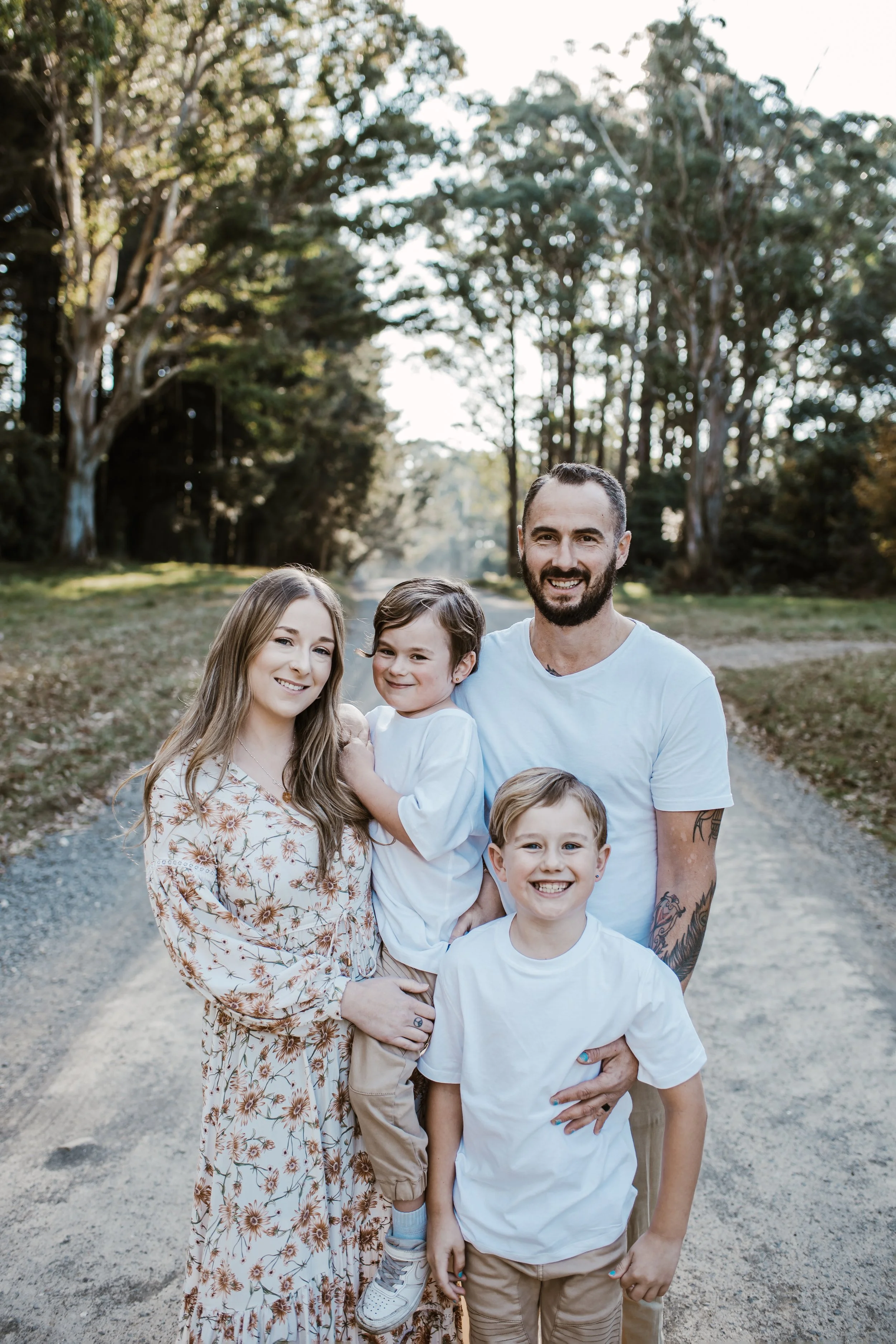 A family of four standing on a dirt path in a park with tall trees in the background. The woman has long blonde hair and is wearing a floral dress. The man has a beard, short dark hair, and tattoos on his arms, and is wearing a white t-shirt. The two children, a girl and a boy, are smiling and dressed in white t-shirts and tan shorts.