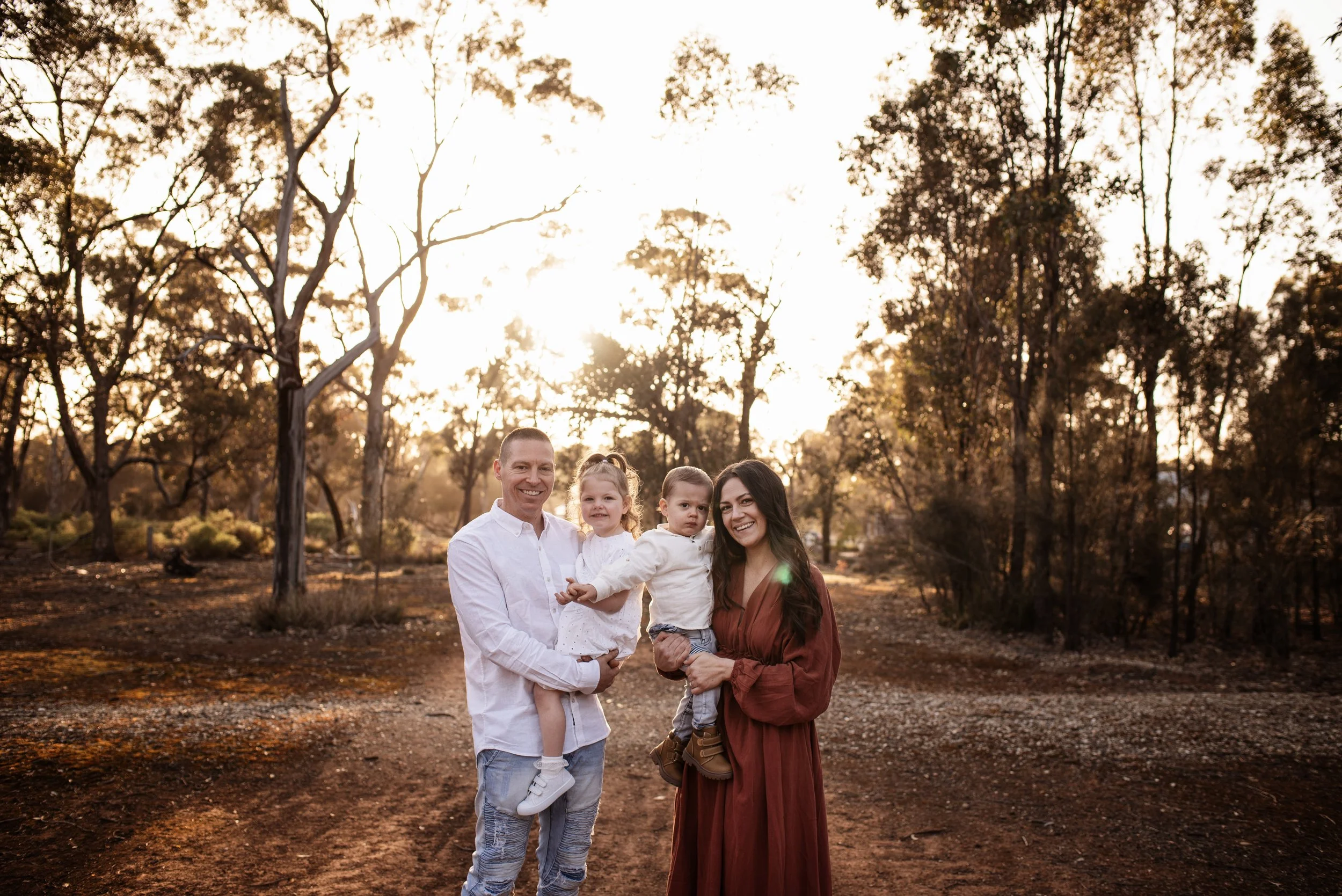 Family of four standing outdoors at sunset in a park-like area with trees in the background. The father, wearing a white shirt, holds a young girl in a white dress. The mother, in a rust-colored dress, holds a young boy in a white shirt and jeans. All are smiling.