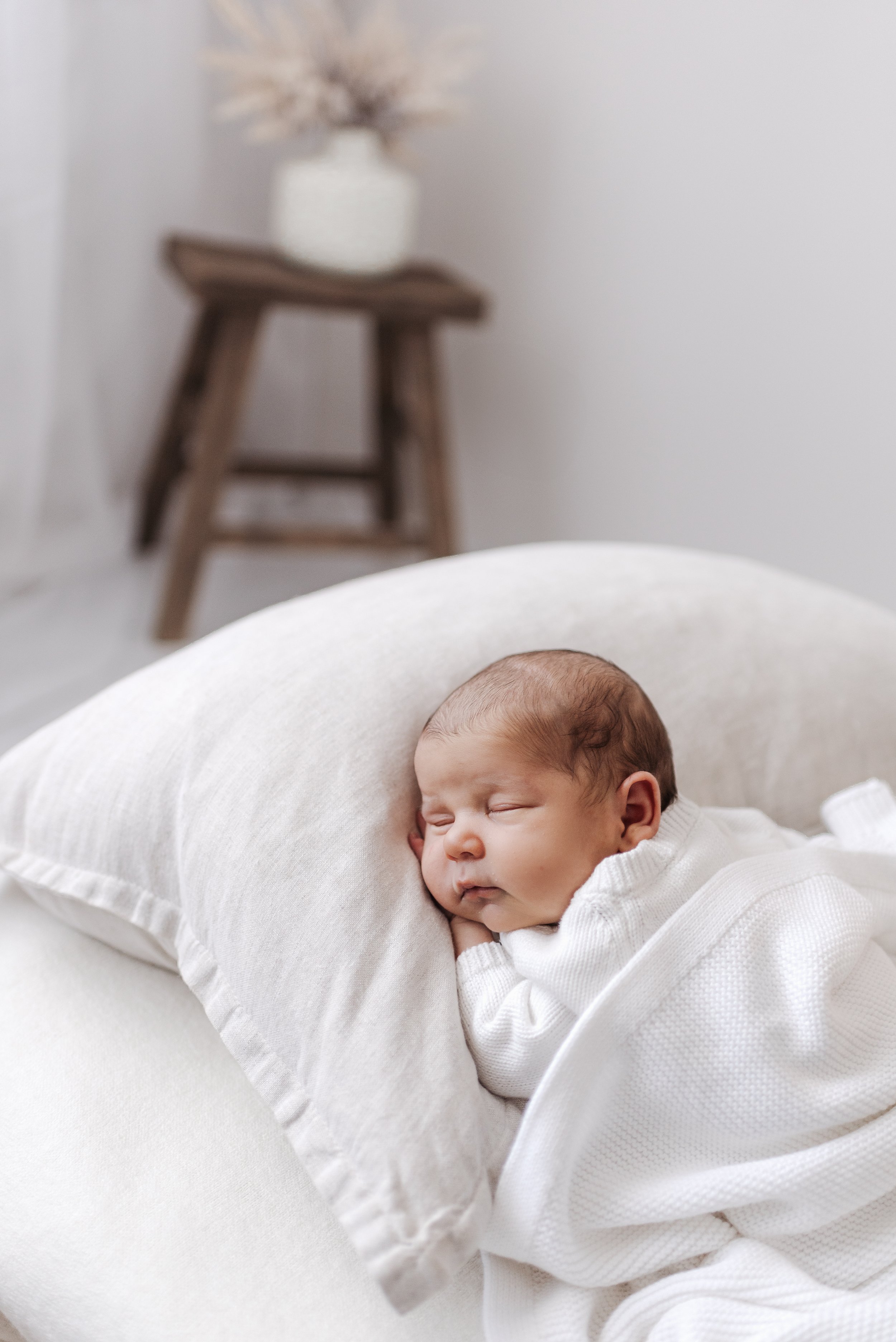 A sleeping newborn baby wearing a white outfit, resting on a white pillow and blanket in a cozy room.