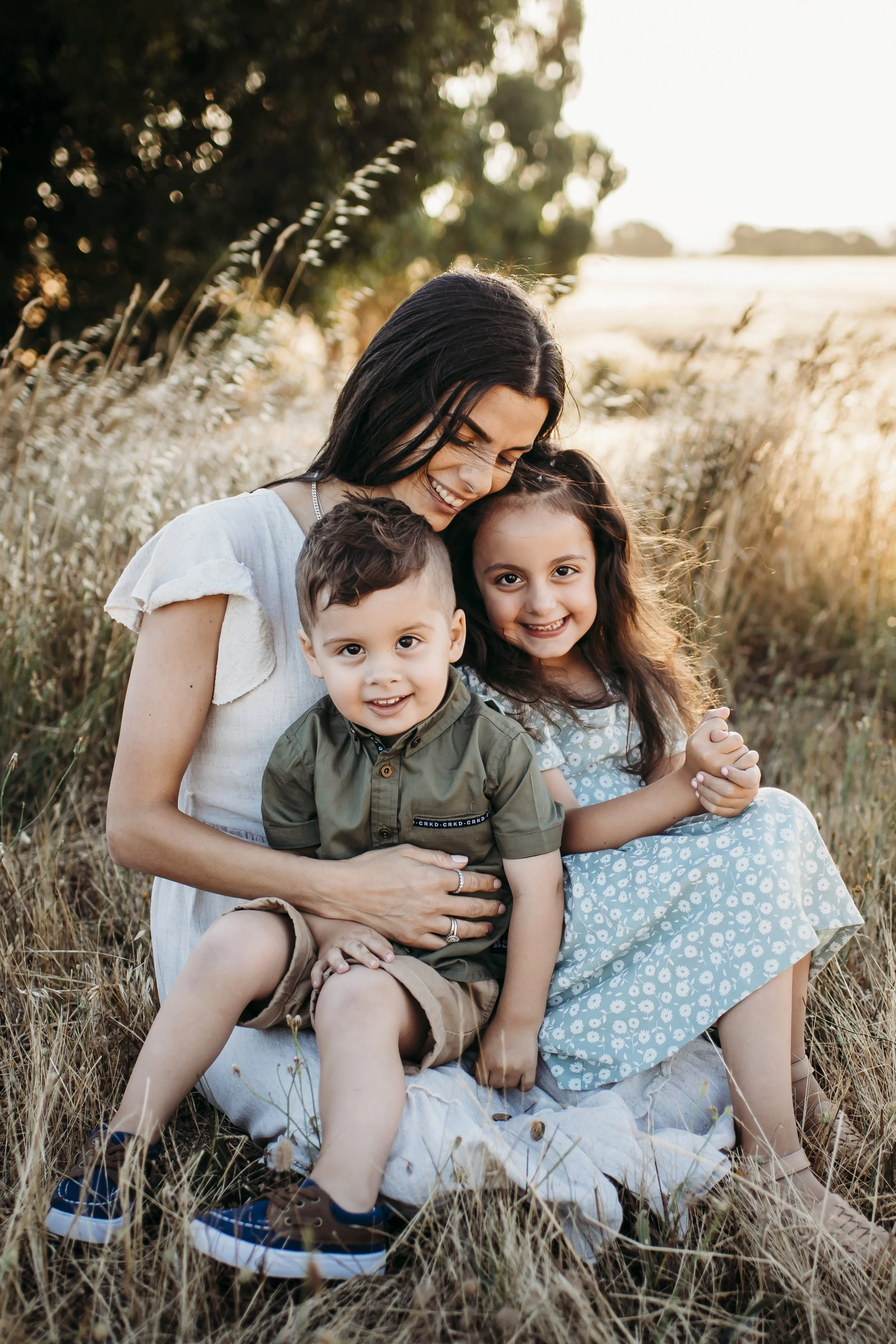 A woman sitting in a field with two children, smiling and hugging each other during sunset.