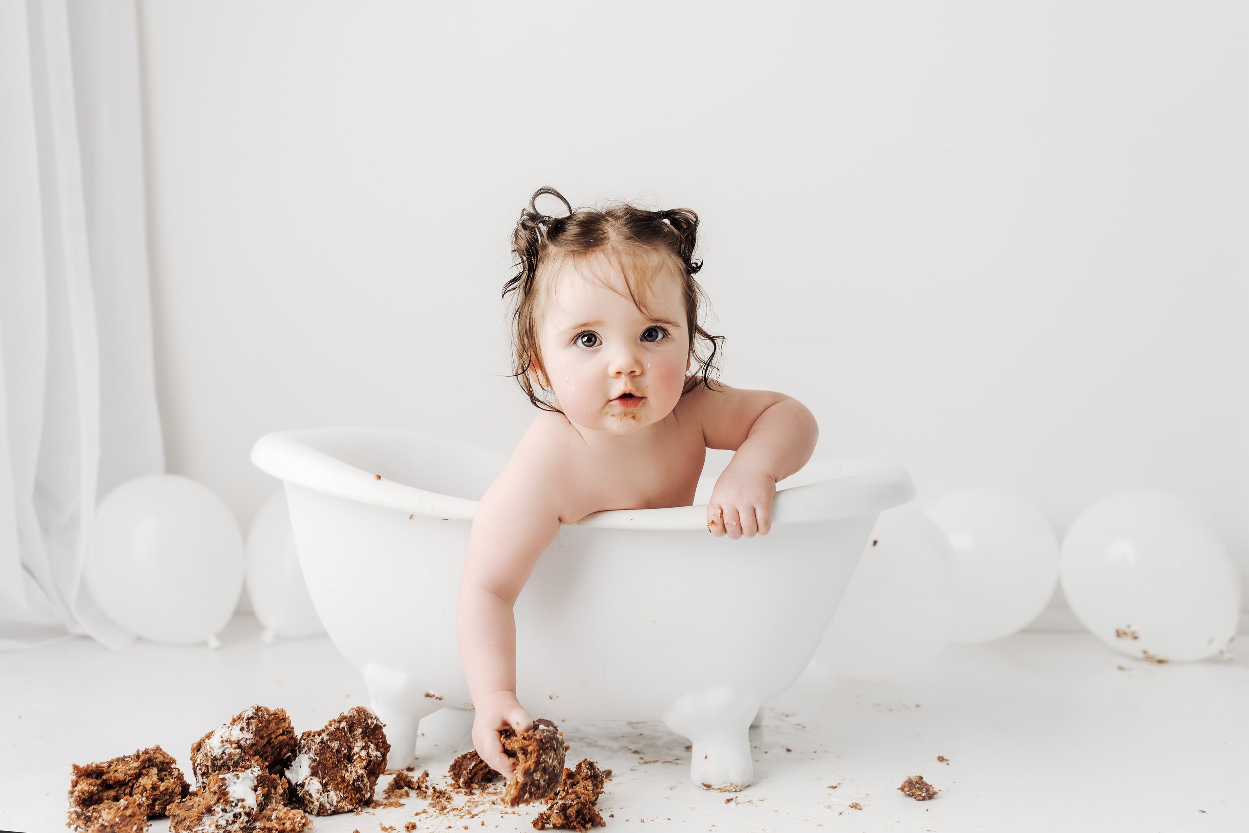A young child with brown hair and big eyes sitting in a white bathtub, surrounded by white balloons and crumbs of cake on the floor and inside the tub, with a surprised expression.