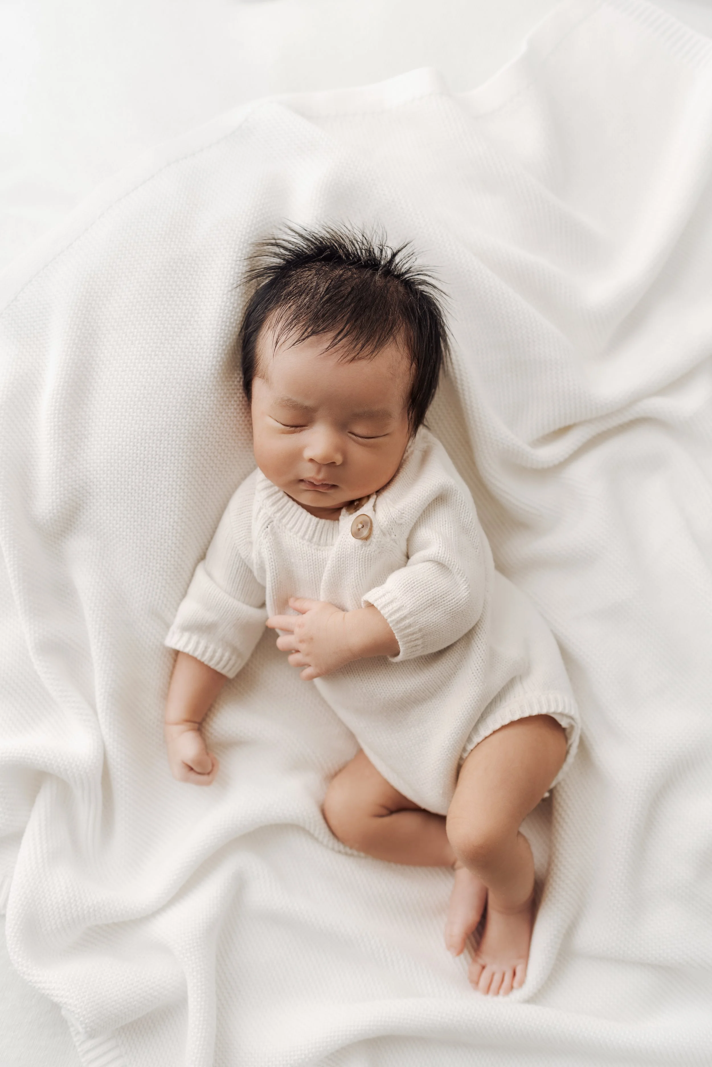 A sleeping infant with dark hair, lying on a white textured blanket, wearing a cream-colored knit outfit.