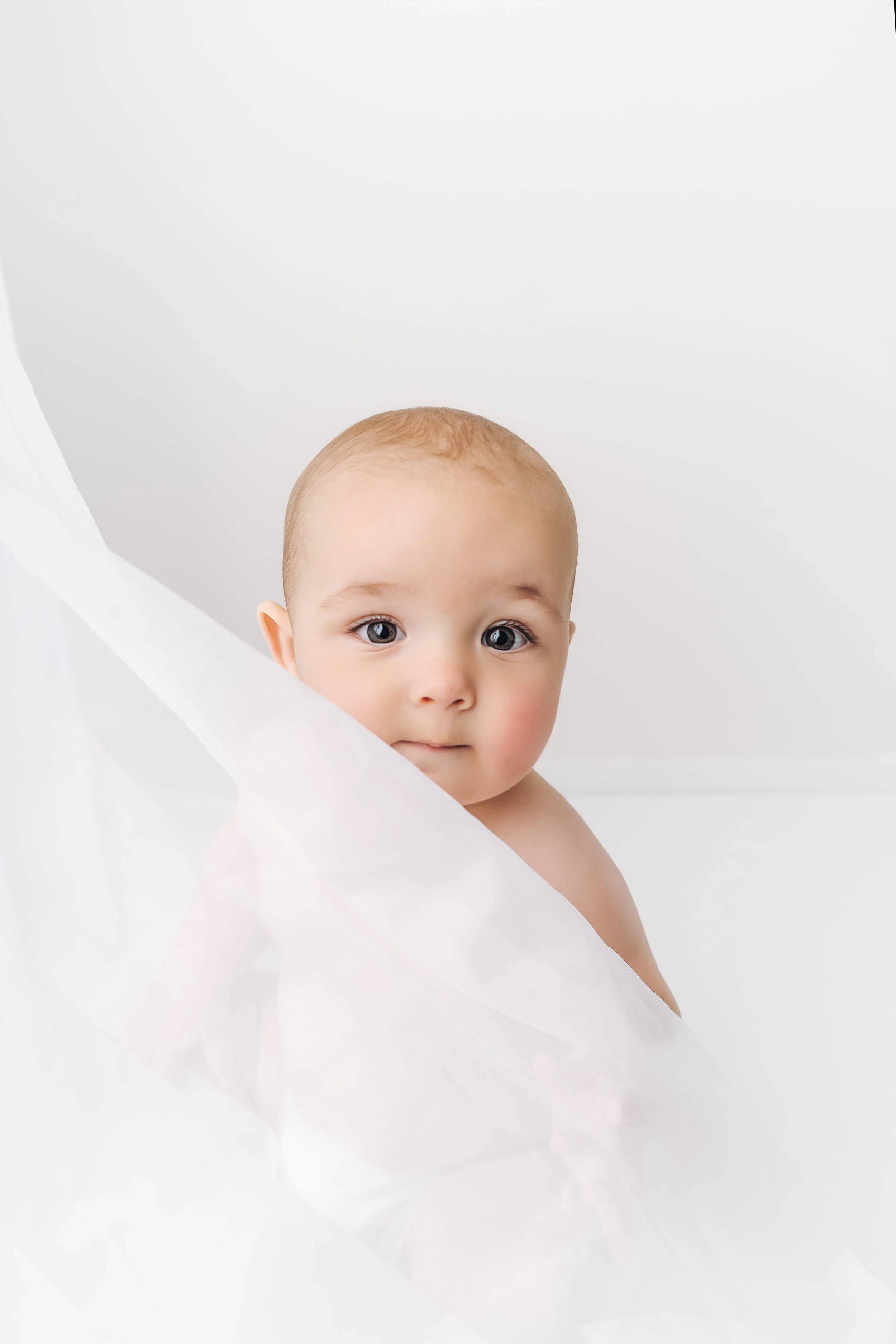 A young baby with blue eyes and light skin peeks from behind a sheer white fabric in a bright, minimal setting.