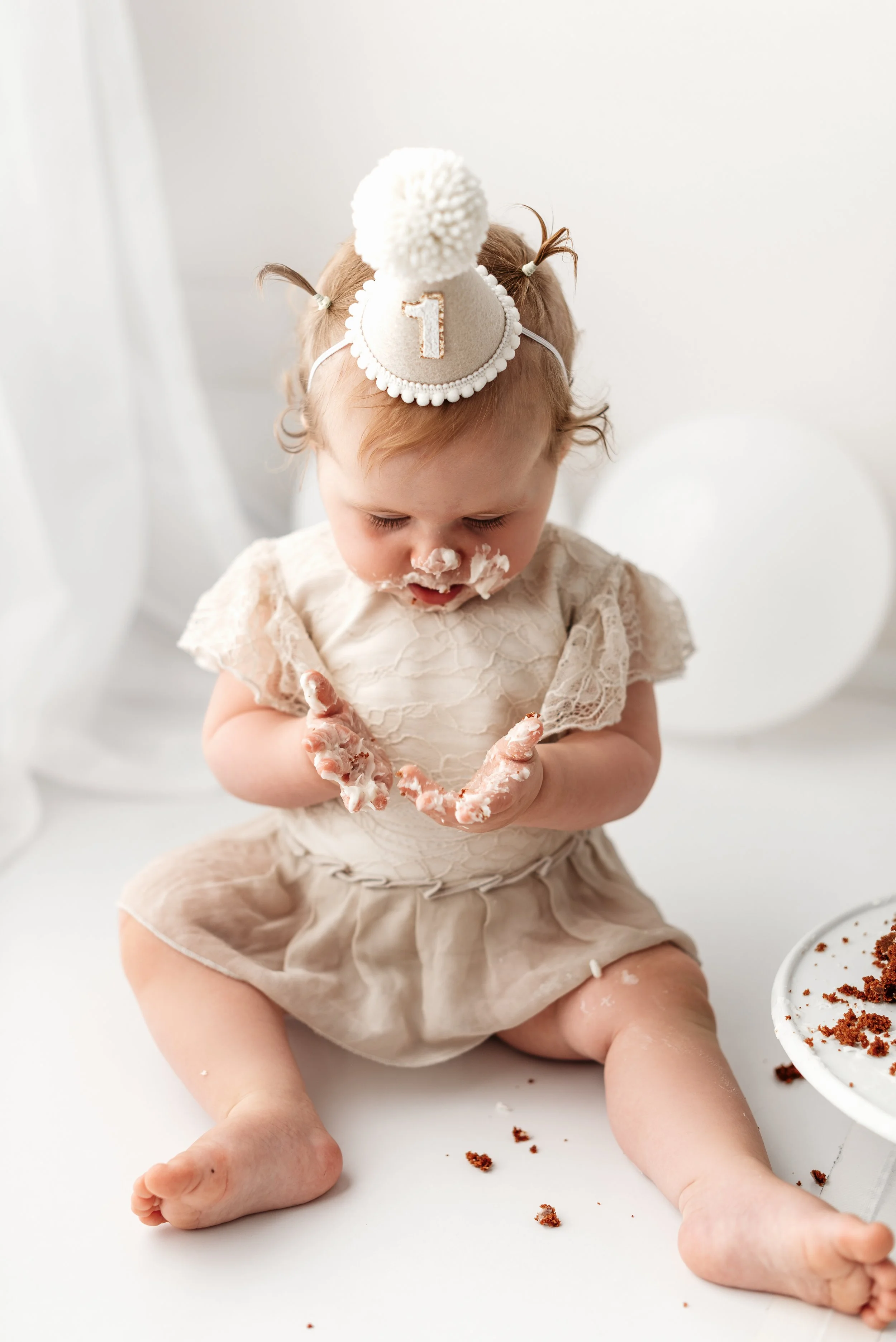 A young child with messy cake and frosting on their face and hands, sitting on the floor with a birthday hat that has a '1' on it, in front of a white background with white balloons.