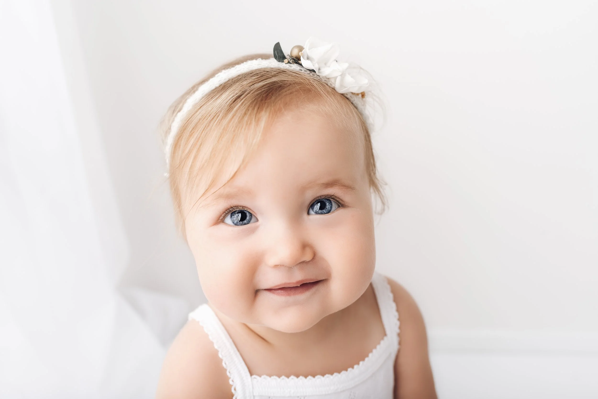 Close-up of a smiling toddler girl with blue eyes, wearing a white headband with flowers, white sleeveless top, and light blonde hair, against a plain white background.