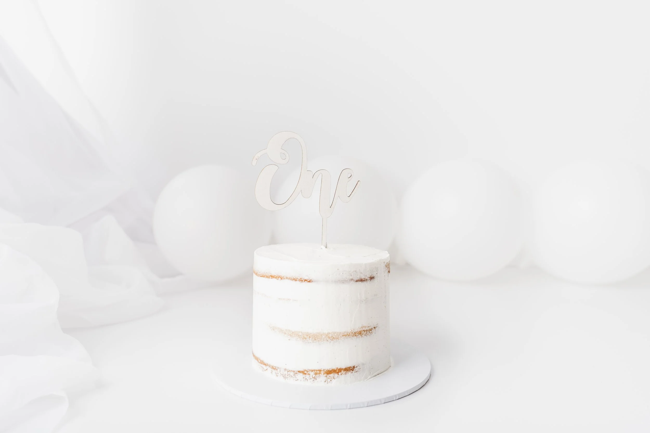 A white wedding cake with minimal decoration and a topper that reads "one" in cursive on a white stand, with white balloons and draped fabric in the background.