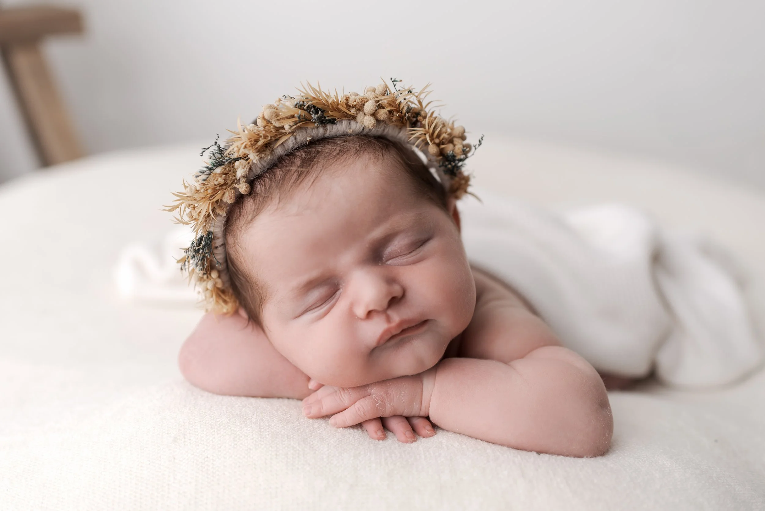 A sleeping baby with a floral crown resting on a soft white surface.