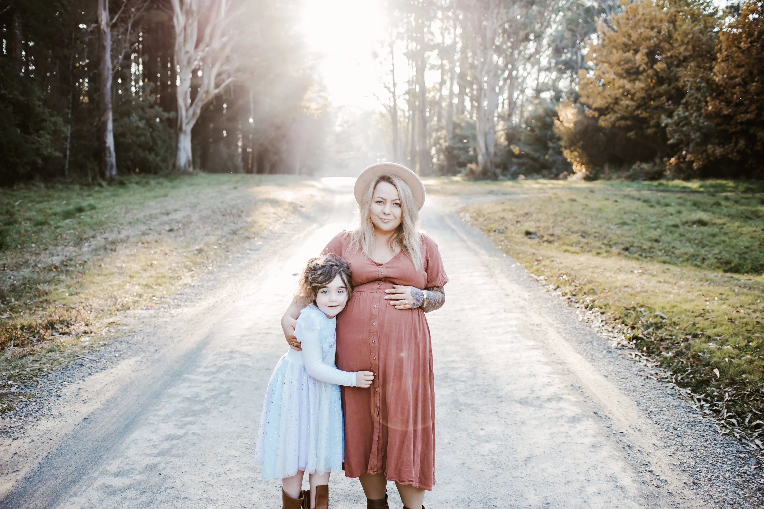 A woman and a young girl standing on a dirt path in a park during sunset, hugging each other. The woman is pregnant, wearing a rust-colored dress and a wide-brimmed hat, and has tattoos on her arm. The girl wears a light blue dress with glittery details and brown boots. There are trees with autumn leaves and sunlight filtering through in the background.
