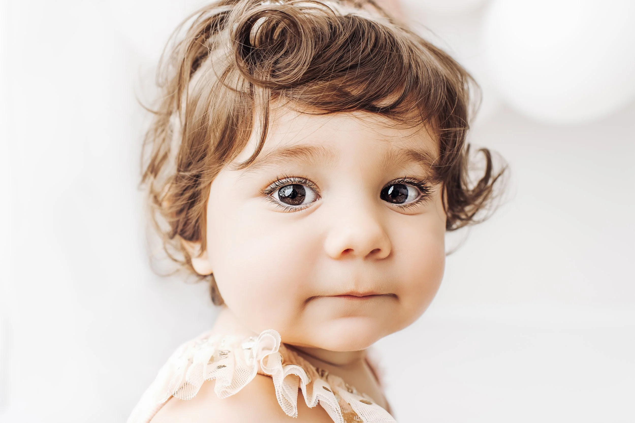 Close-up of a young child with curly brown hair, big blue eyes, and a soft expression, wearing a beige dress with ruffled lace shoulders.