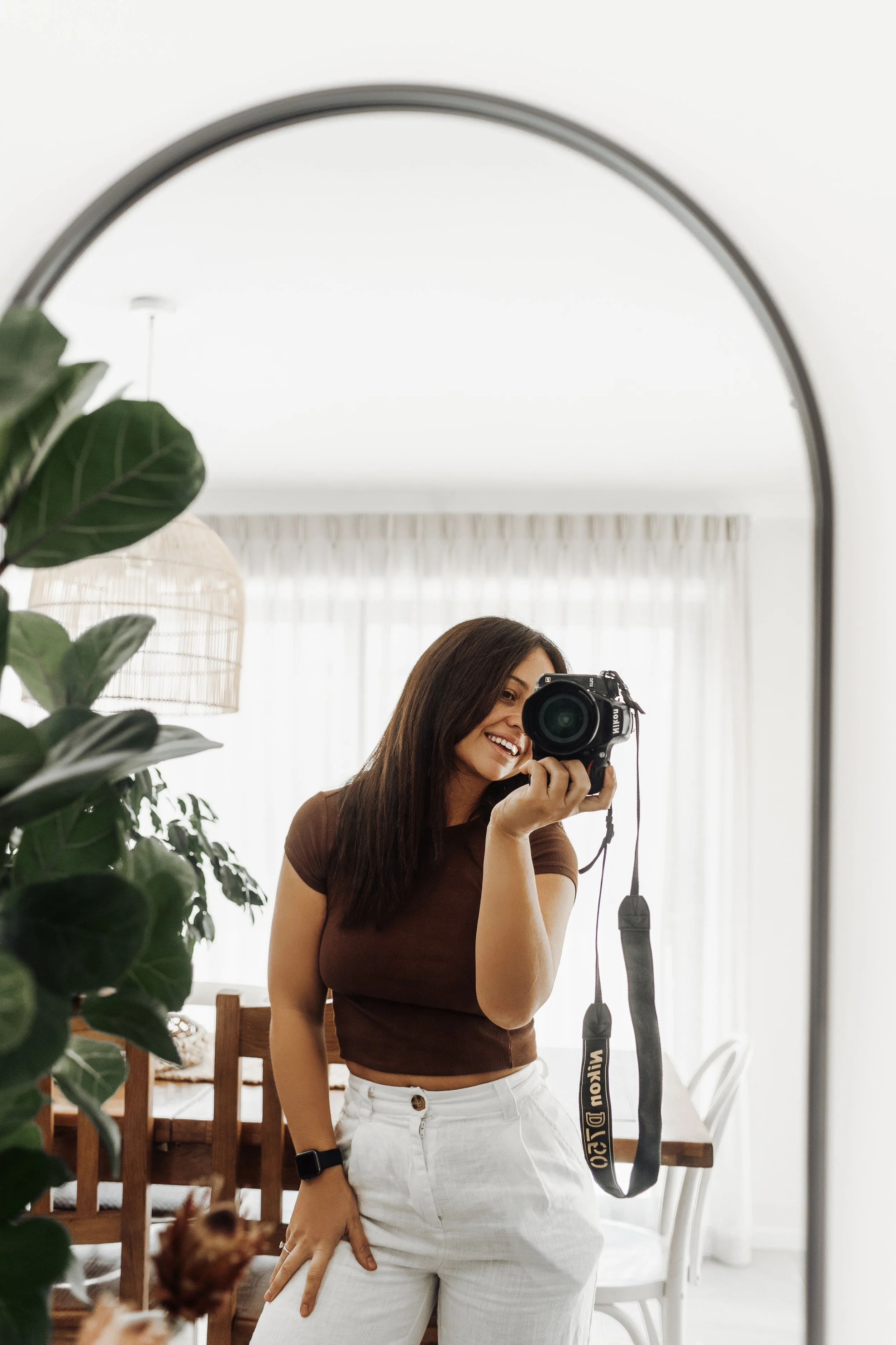 A woman smiling and taking a selfie with a camera in front of a mirror in a bright, modern home with plants, a dining table, and sheer curtains.