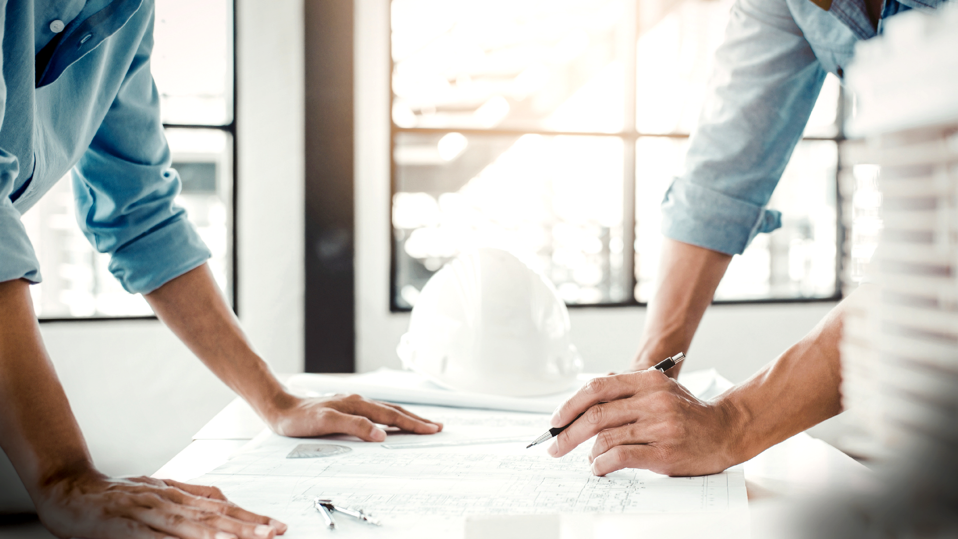 Two individuals are working on architectural plans at a desk in a well-lit office. One is holding a pen and pointing at the plans, while the other has hands resting on the table. A white hard hat and large windows are visible in the background.