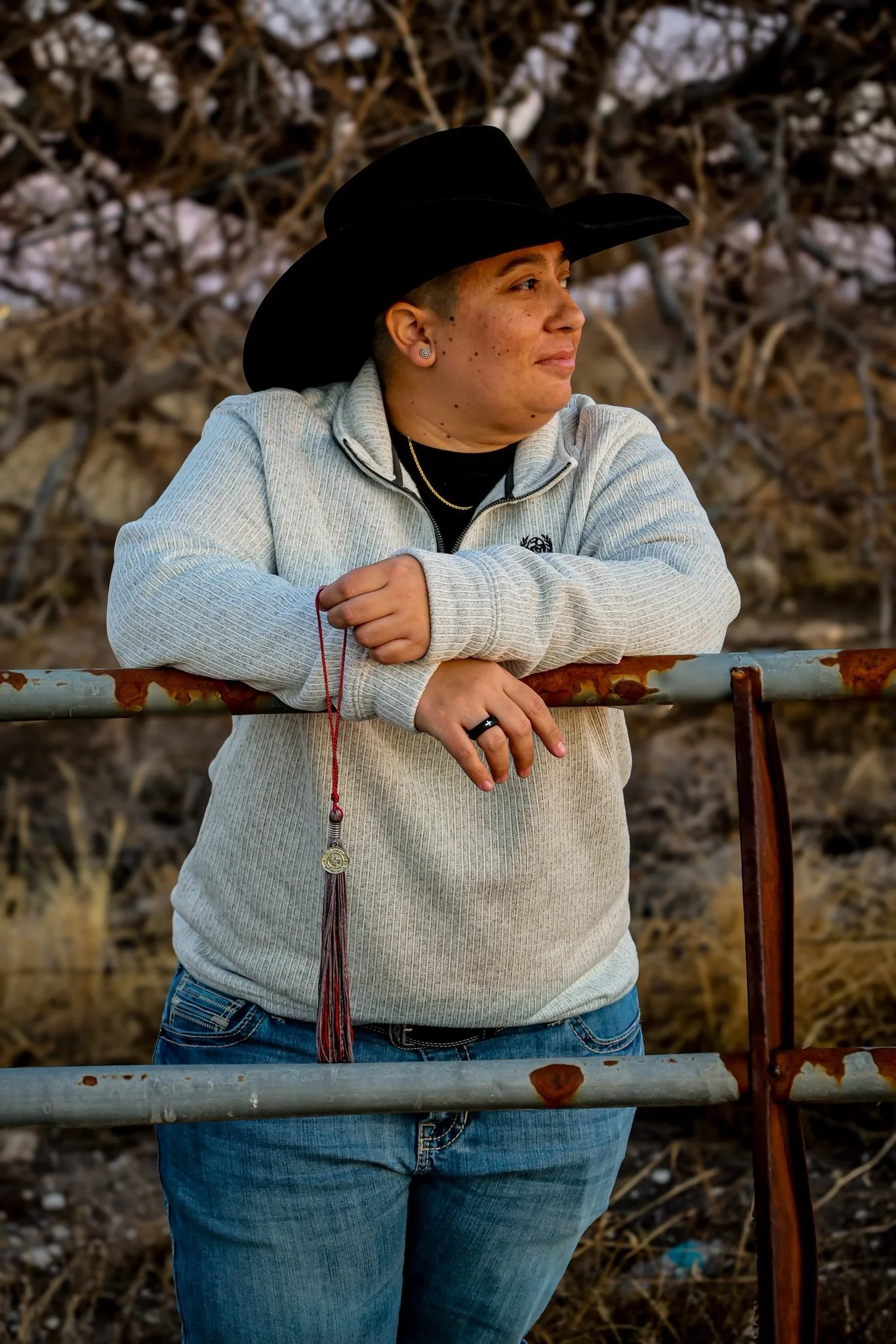 A woman wearing a black cowboy hat and a light gray North Face sweatshirt leaning on a rusted metal gate outdoors in a natural setting with dry trees and grass.