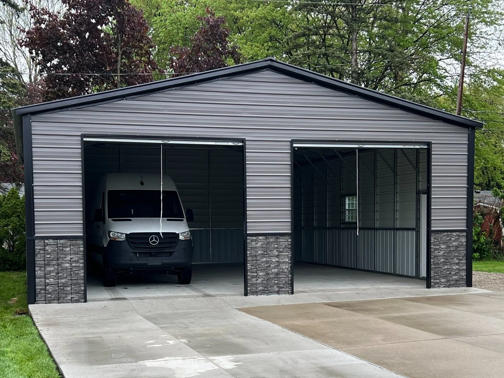 New metal garage with two bays, one with a Mercedes-Benz van inside, concrete driveway, and green trees in the background.