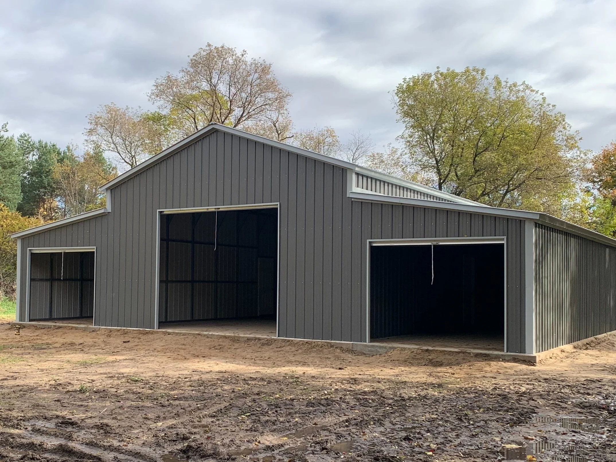 Gray metal storage building under construction with open bays, surrounded by trees with fall foliage and mud ground.