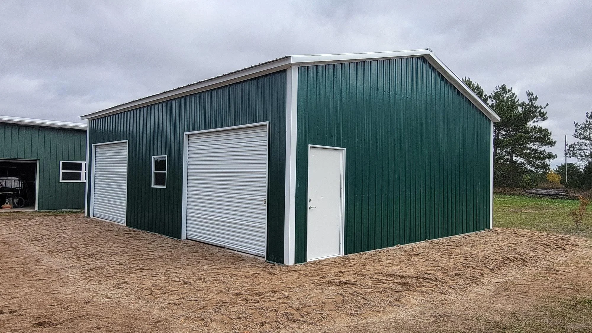 Green metal building with two white roll-up garage doors, a white side door, and two small windows, located on a dirt lot under a cloudy sky.
