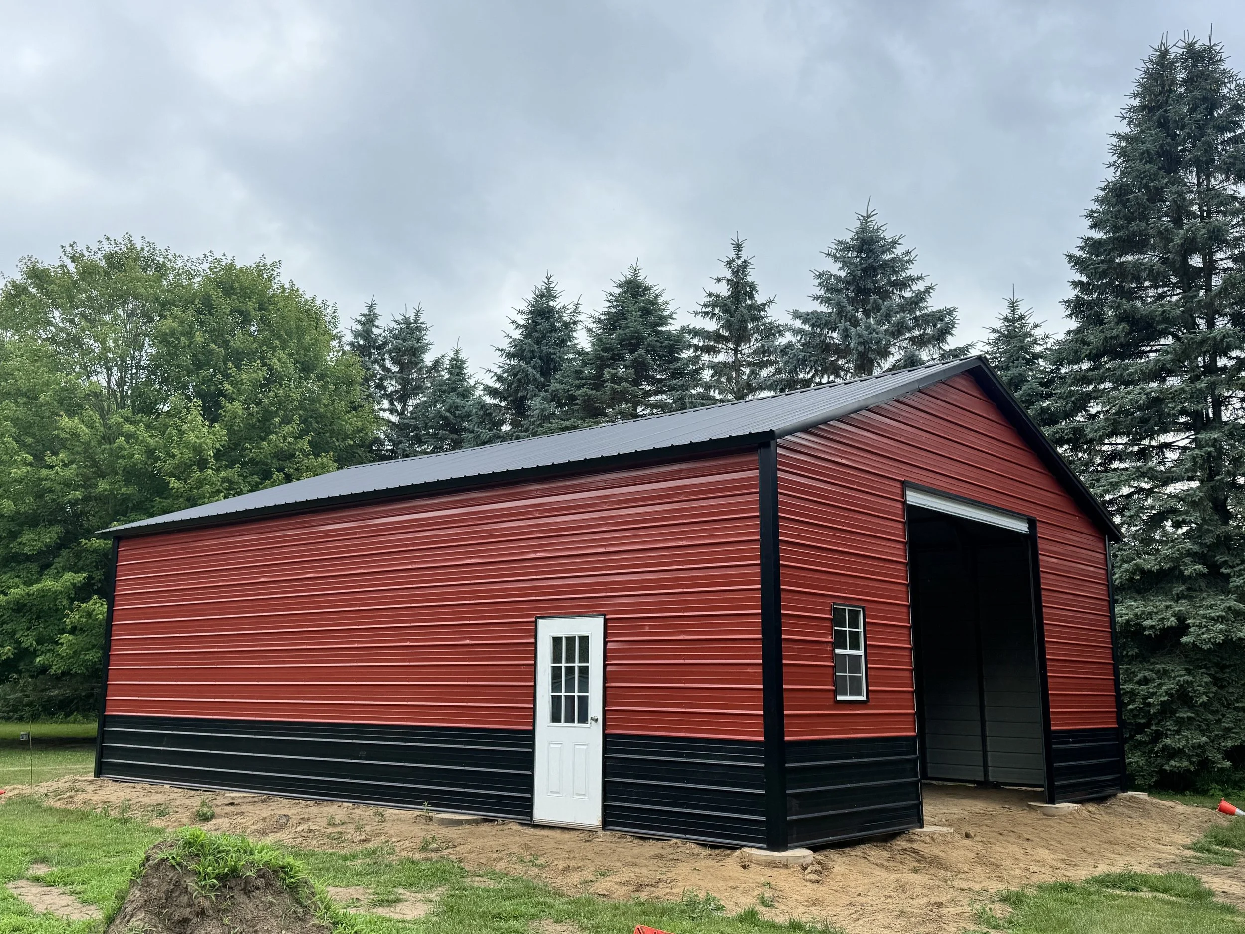Red and black metal barn with a white door and small window, situated on a patch of dirt with a grassy area, surrounded by green trees and a cloudy sky.