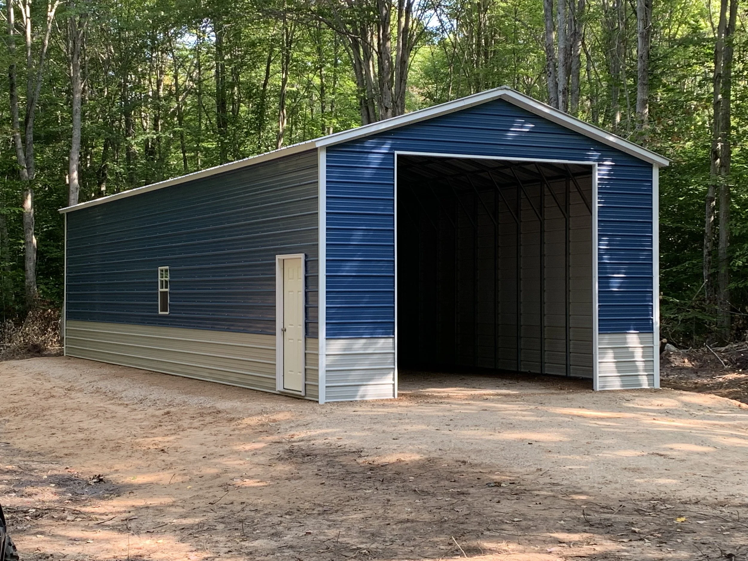 A large metal storage building with blue and gray siding, a small side door, and open entryway, situated in a wooded area on a dirt surface.