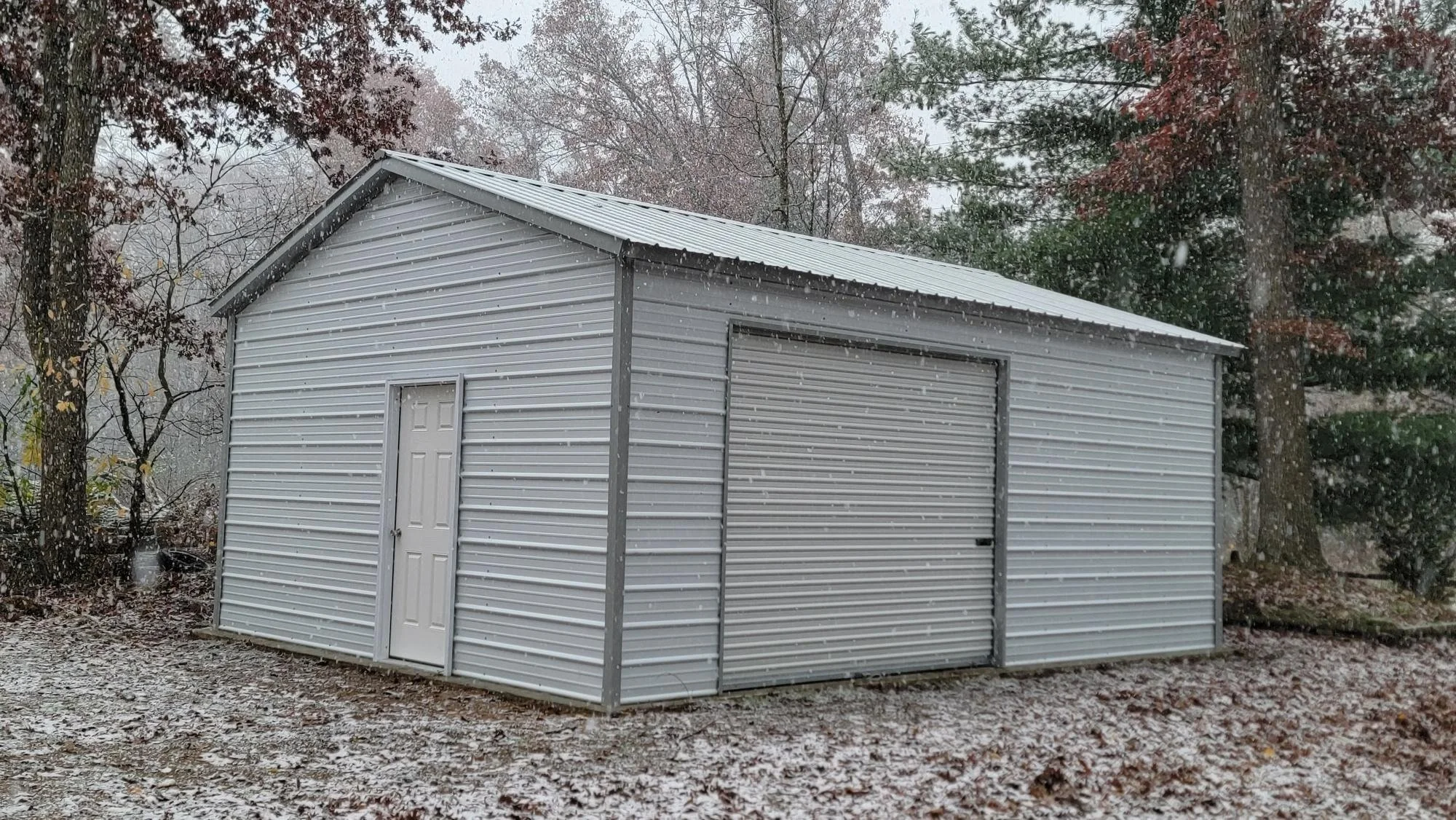 A white metal shed with a gabled roof and two doors, located outdoors among trees with autumn-colored leaves, during a snowfall.