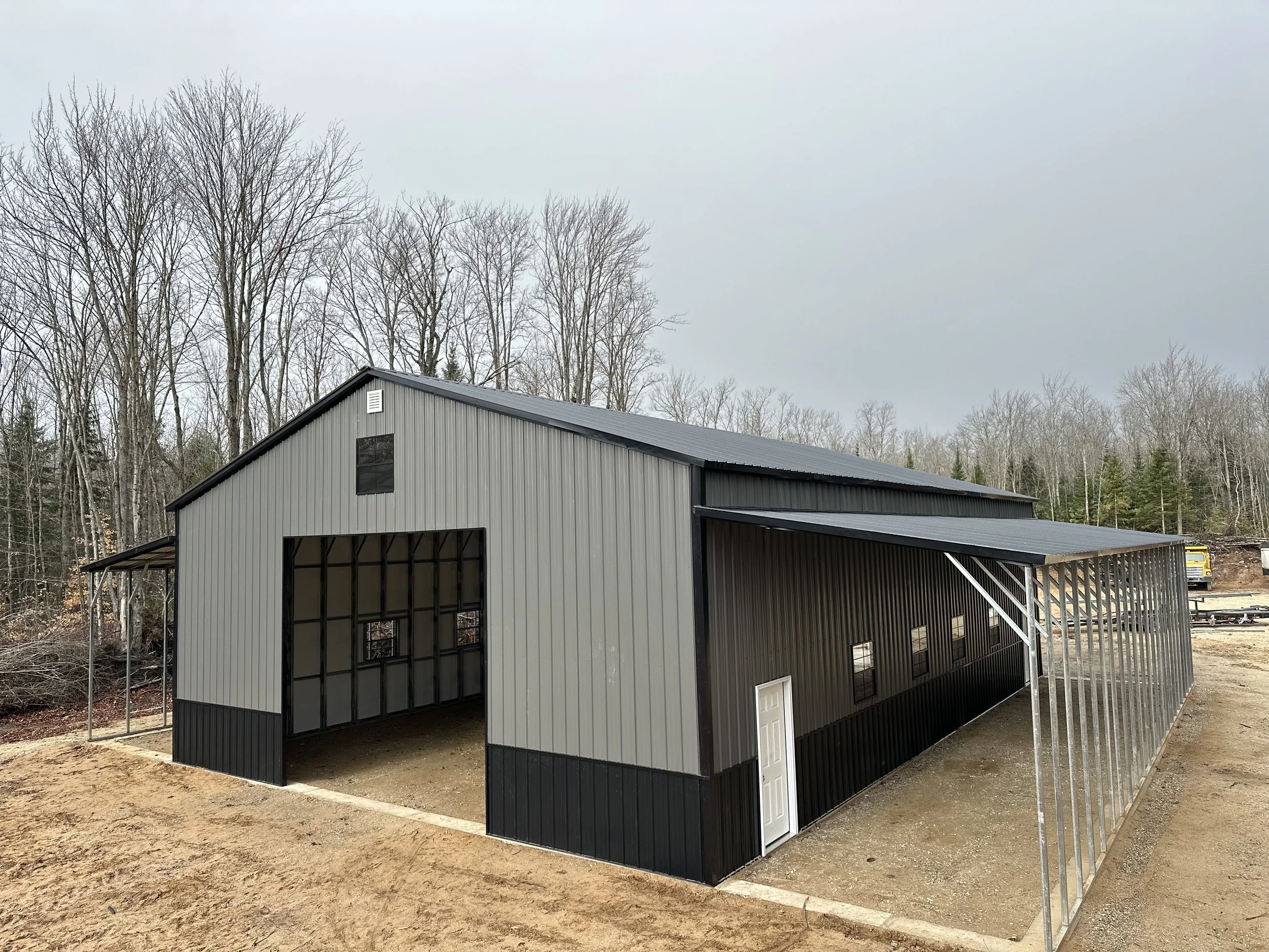 Unfinished metal building structure with gray and black siding and a sloped roof, surrounded by bare trees and construction materials.
