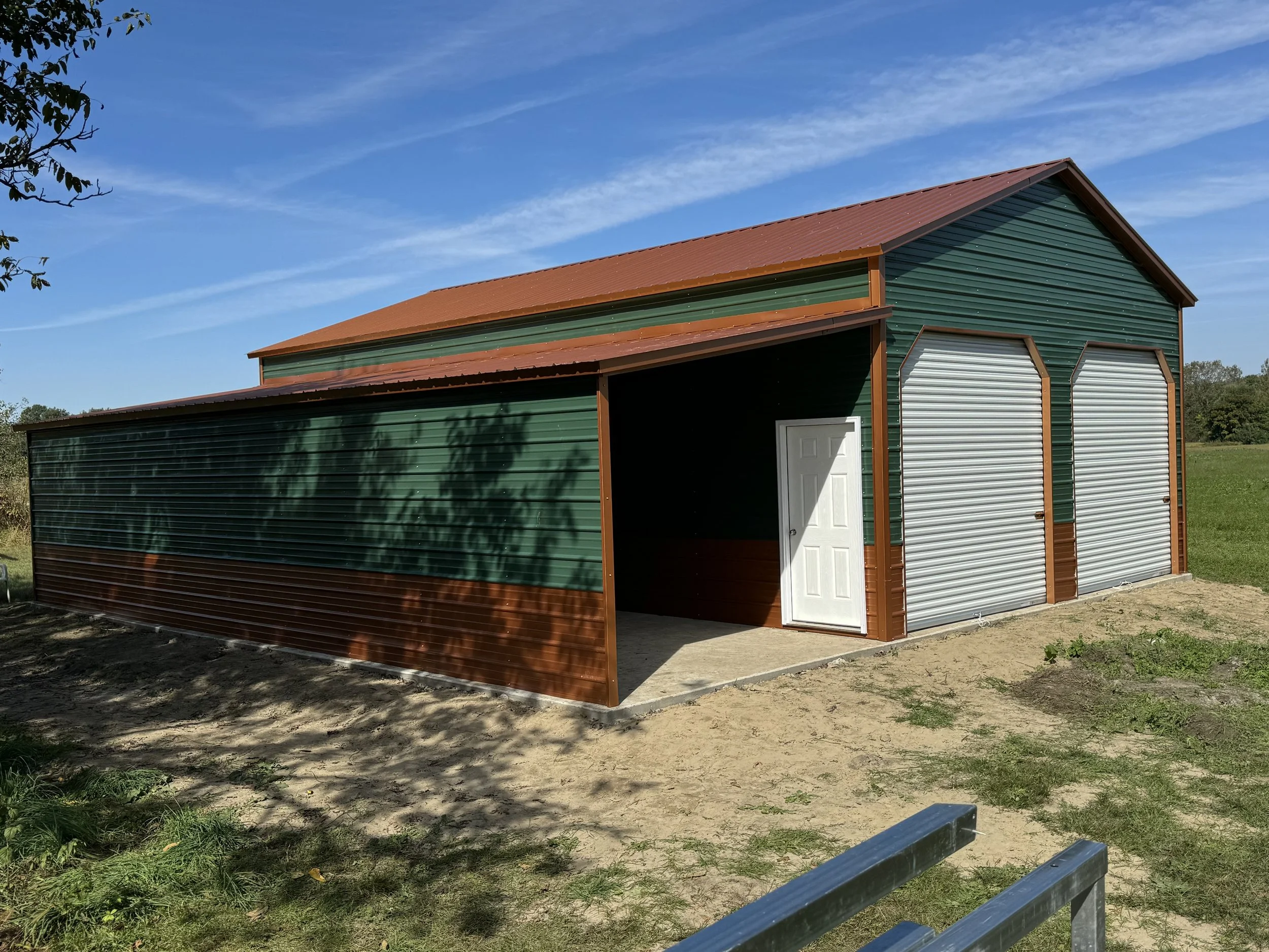 Green barn with brown trim and a red metal roof, featuring two large white roll-up doors and a side door, situated on a grassy field with a clear blue sky overhead.